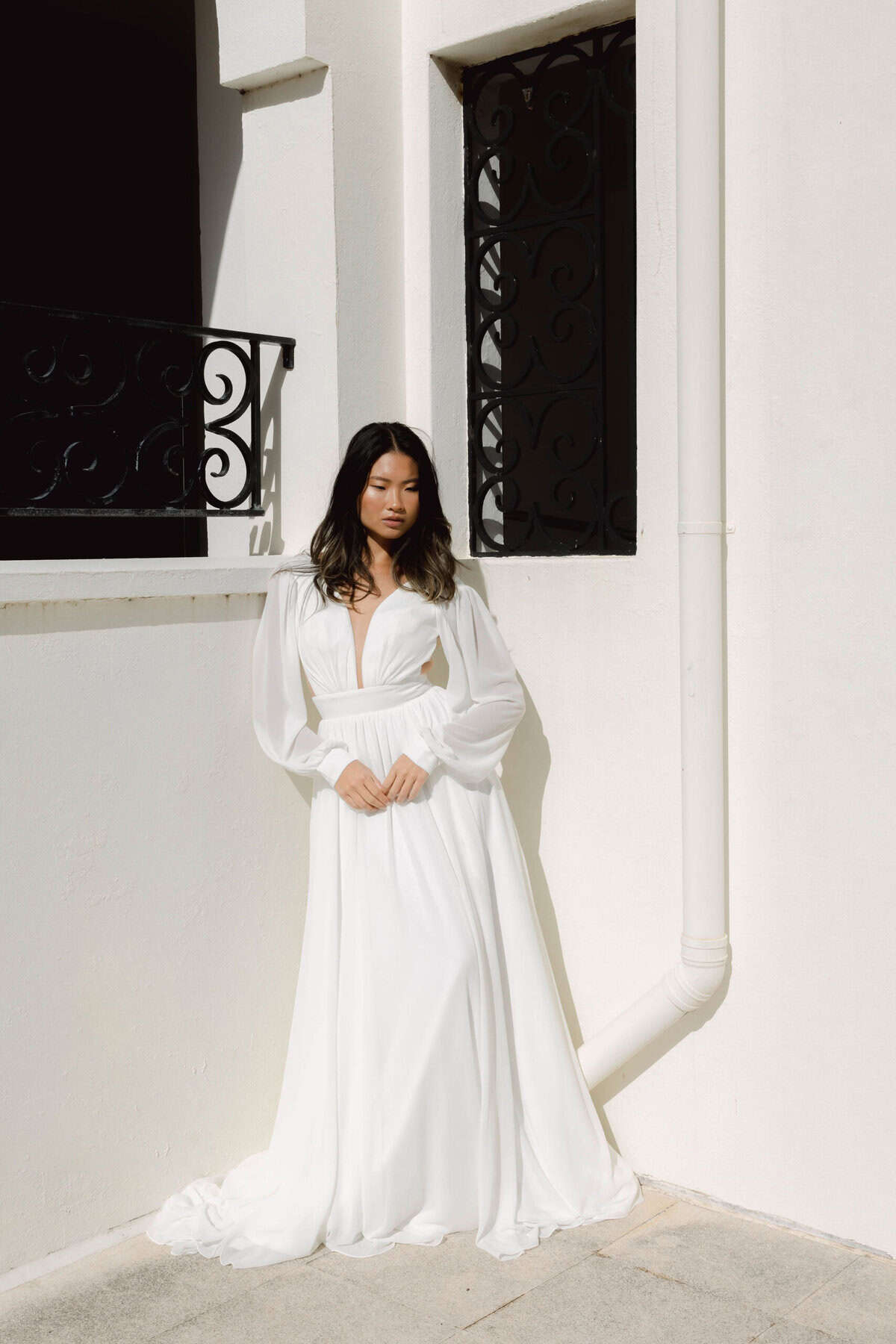 Beautiful Asian bride in a flowing white wedding dress standing against a minimalist white building with black window details.