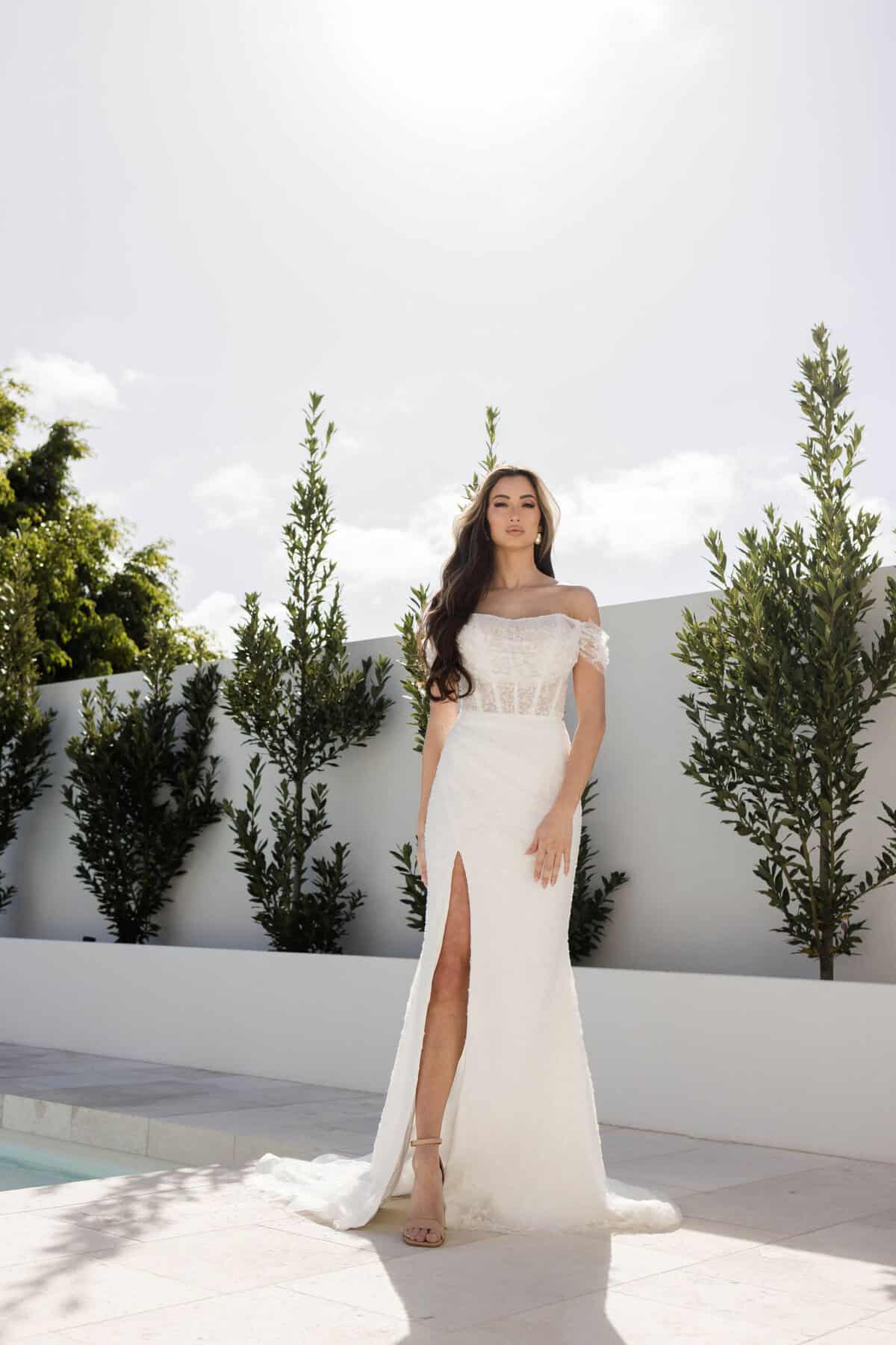 A woman in a glamorous bridal gown with an off-shoulder design and thigh-high slit stands by a pool, surrounded by greenery and a white wall, under a bright, sunny sky.