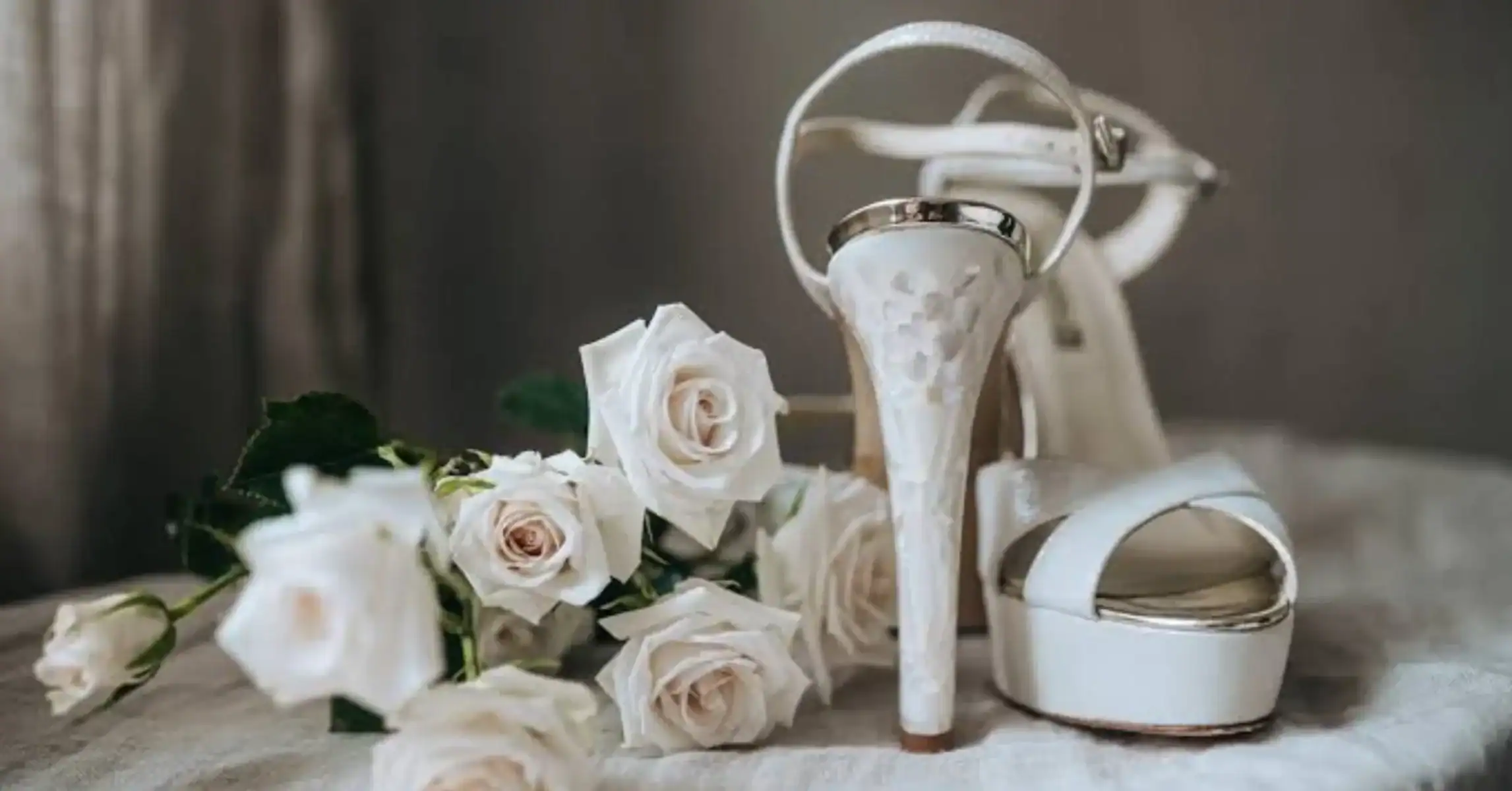 A pair of white high-heeled wedding shoes in Singapore with gold accents is placed beside a bouquet of white roses on a soft, light-coloured surface, evoking elegance for a special occasion or wedding.