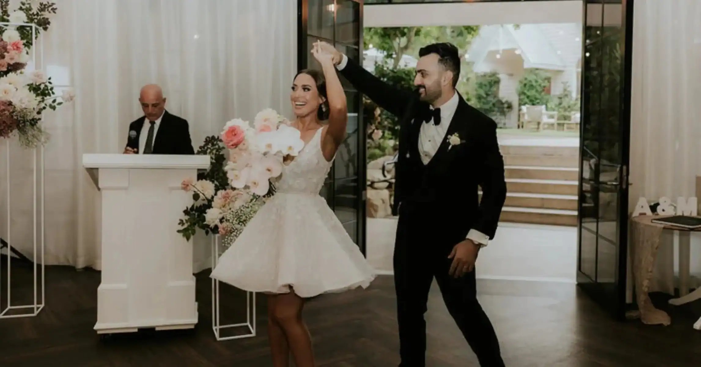 Radiant and stylish, this bride shows What to Wear for Your Wedding Dinner in a chic short white dress, holding a bouquet as she enters hand-in-hand with her groom in a black tuxedo. Flowers and elegance fill the room.