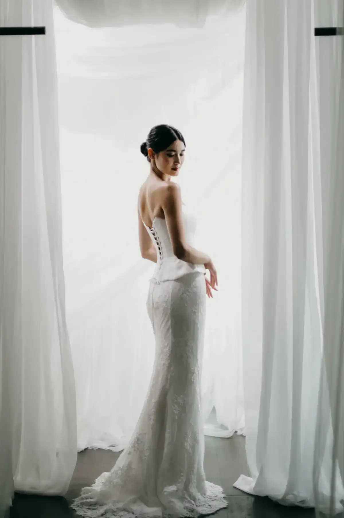 A woman in a strapless white lace wedding dress stands gracefully between sheer white curtains, looking over her shoulder in soft natural light.