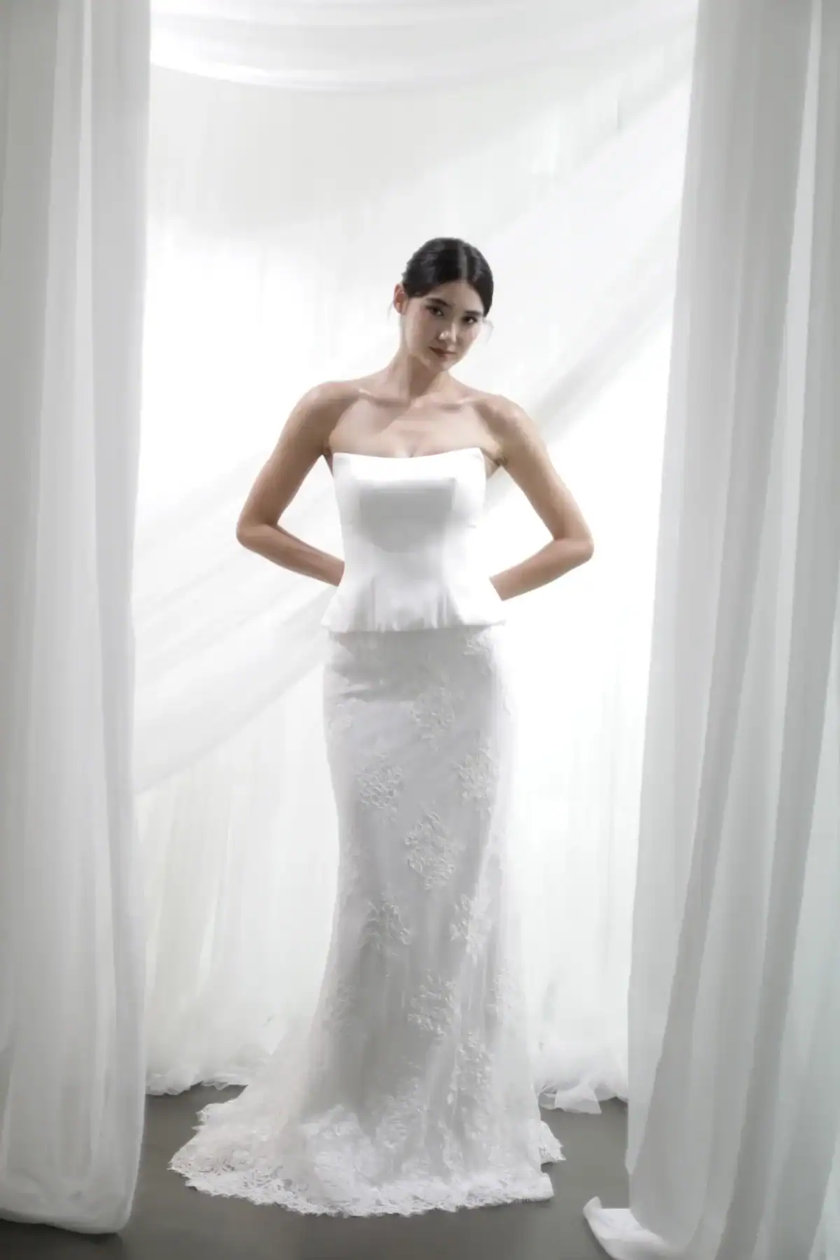 A woman in a strapless white wedding dress with lace details stands between sheer white curtains in a softly lit room, looking slightly to the side with her hands on her hips.