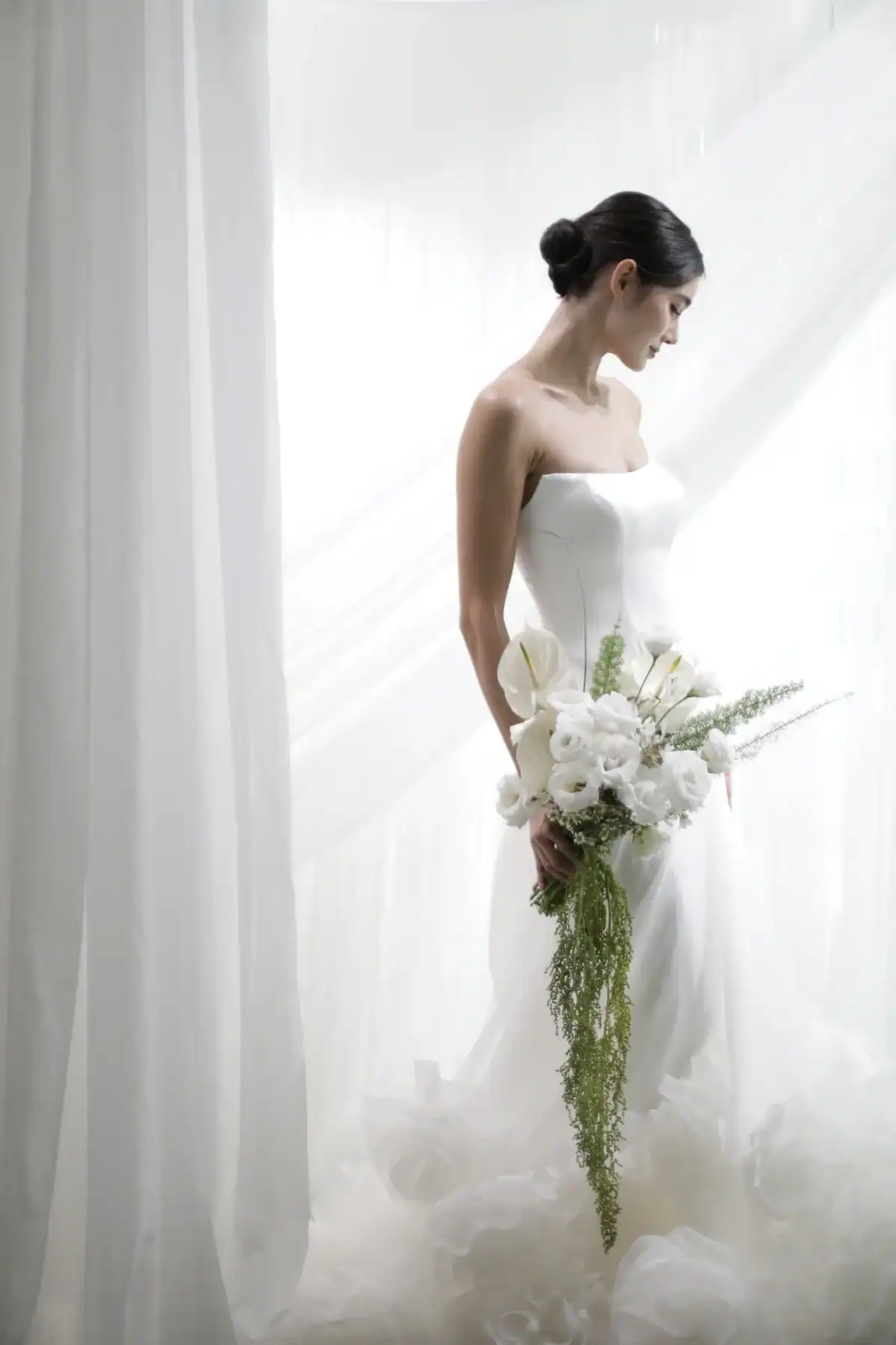 A bride in a strapless white wedding dress stands by sheer white curtains, holding a bouquet of white flowers and greenery, looking down with her hair styled in a low bun.