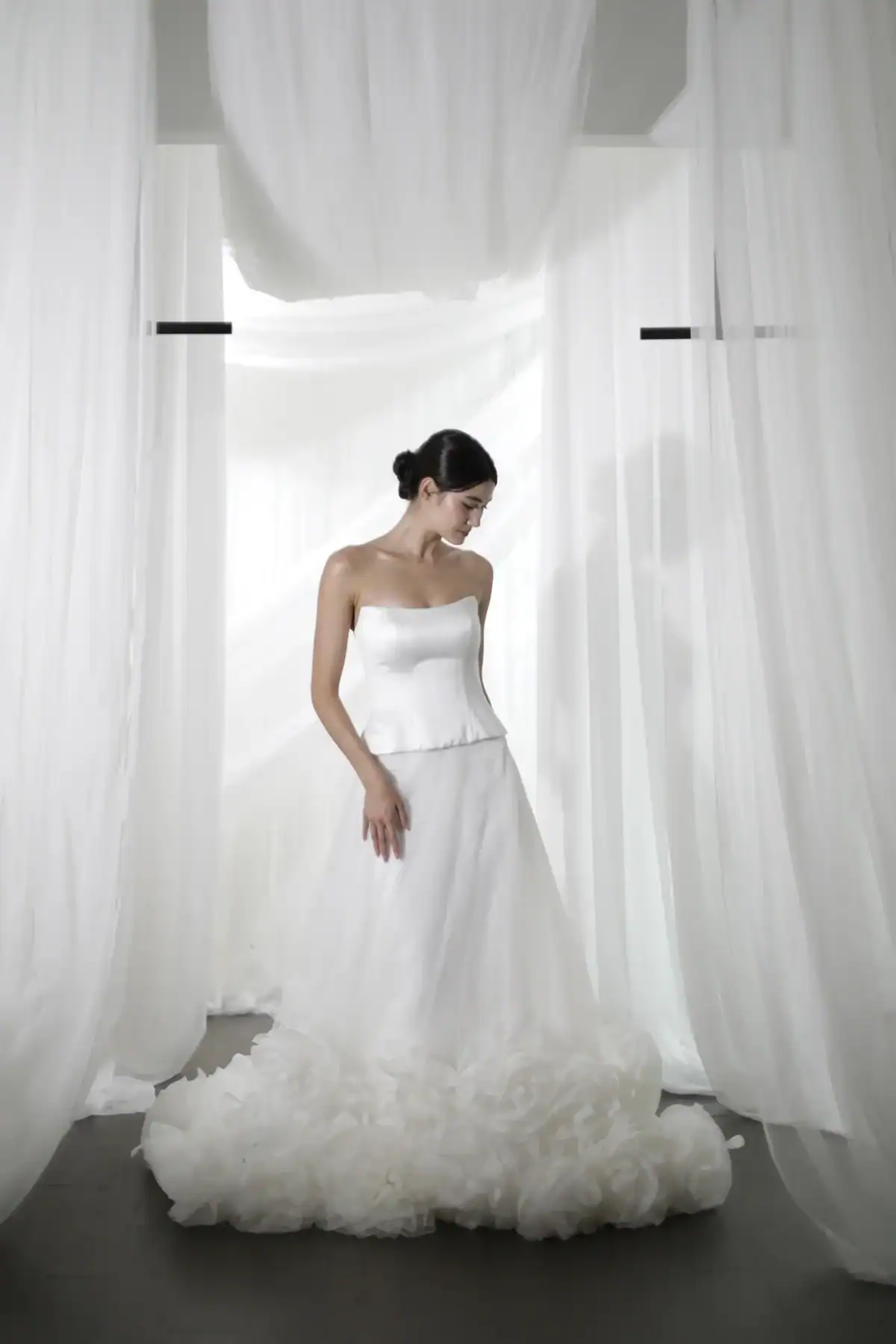 A woman in a strapless white wedding dress stands among flowing white curtains, looking down with her hair styled in a low bun. The dress features a fitted bodice and a voluminous, textured skirt.