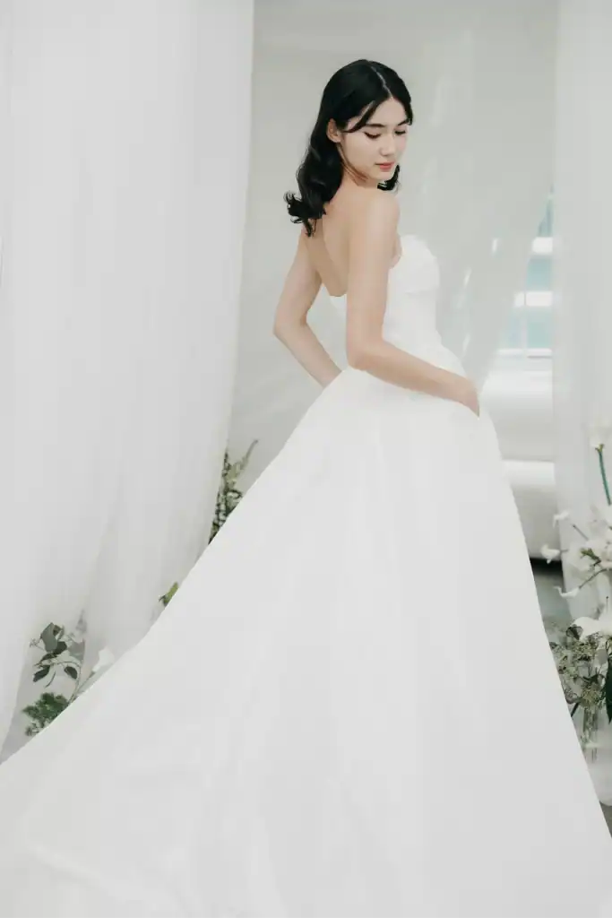 A woman with dark hair stands in a white strapless wedding dress, looking over her shoulder. She is surrounded by soft white curtains and greenery, creating an elegant and serene atmosphere.