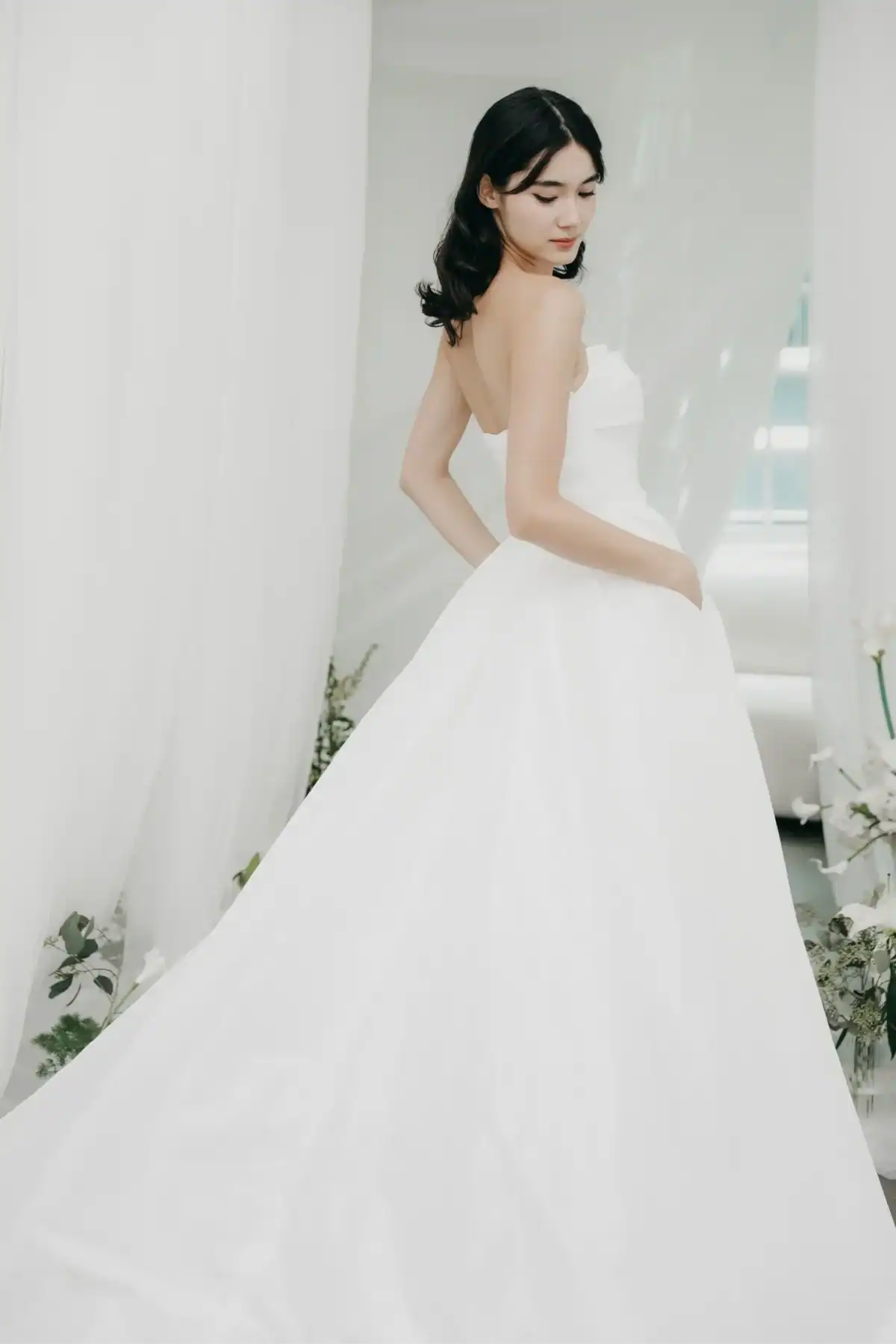 A woman with dark hair stands in a white strapless wedding dress, looking over her shoulder. She is surrounded by soft white curtains and greenery, creating an elegant and serene atmosphere.