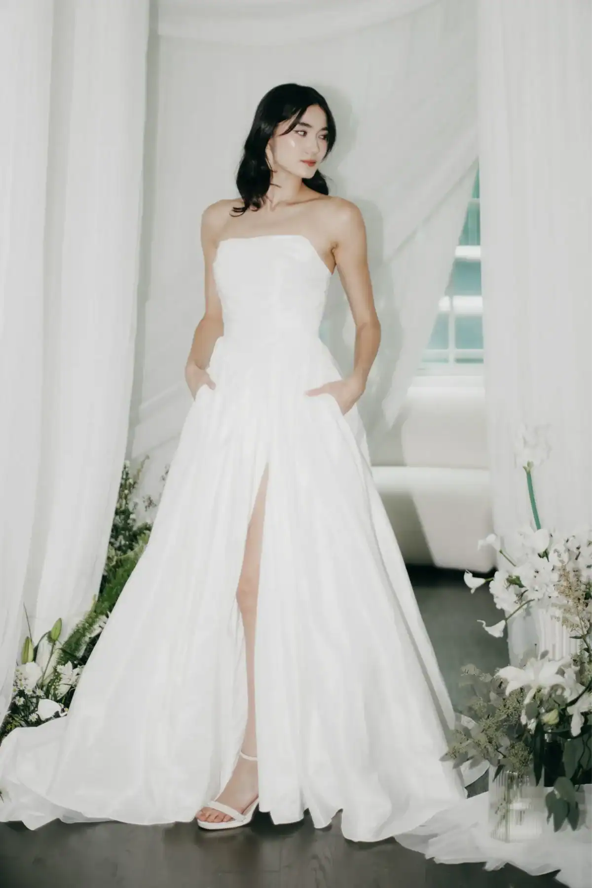 A woman in a strapless white wedding dress with a thigh-high slit stands indoors among white drapes and floral arrangements, looking to the side with her hands in her dress pockets.