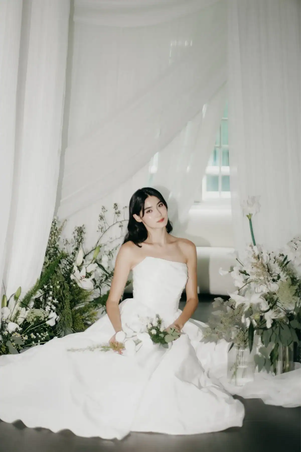 A woman in a strapless white gown sits gracefully among white flowers and greenery, with sheer white drapery hanging behind her and soft natural light filtering in from a window.