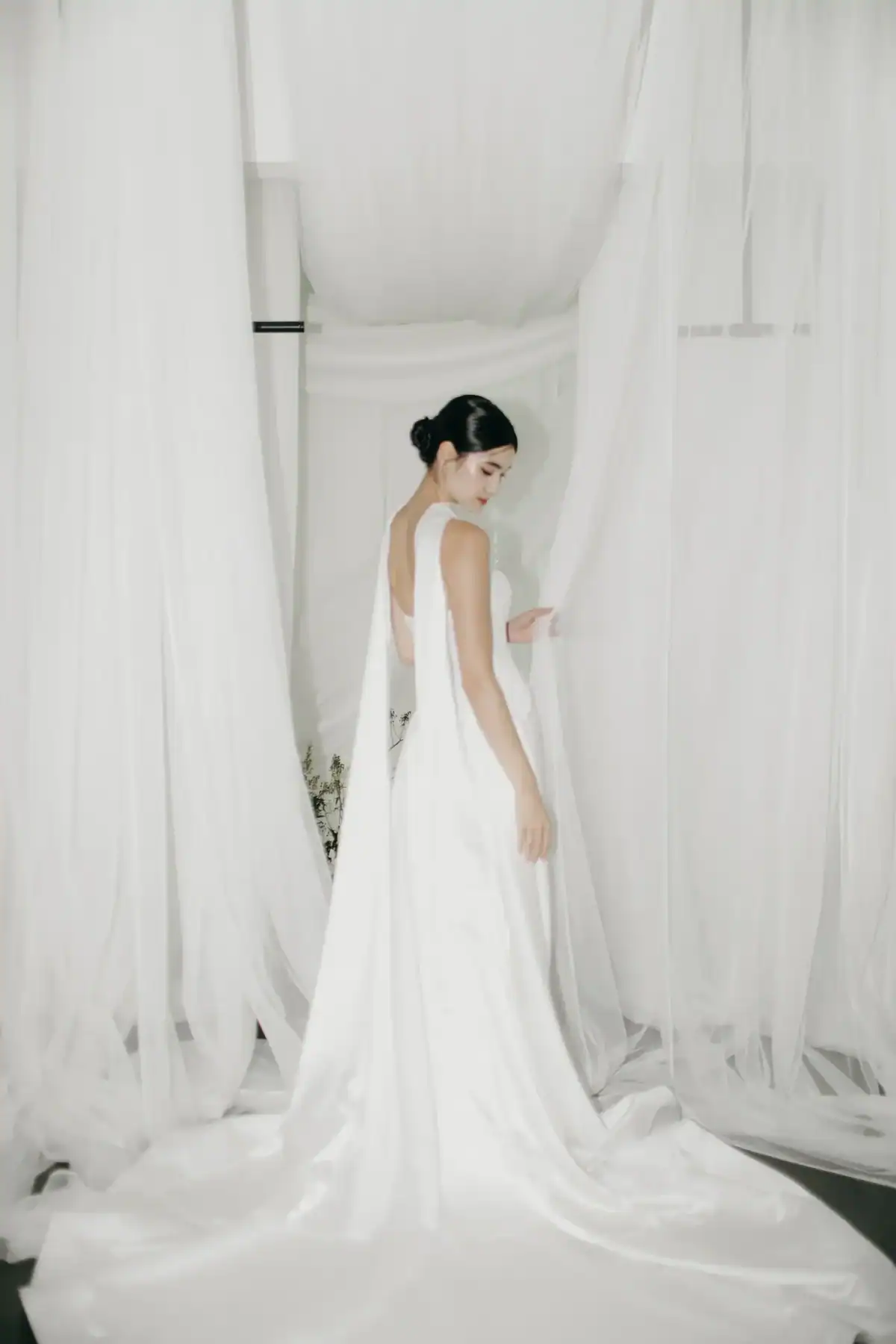 A woman in a white, sleeveless, backless wedding dress stands gracefully among sheer white curtains, looking over her shoulder with her hair styled in a low bun.