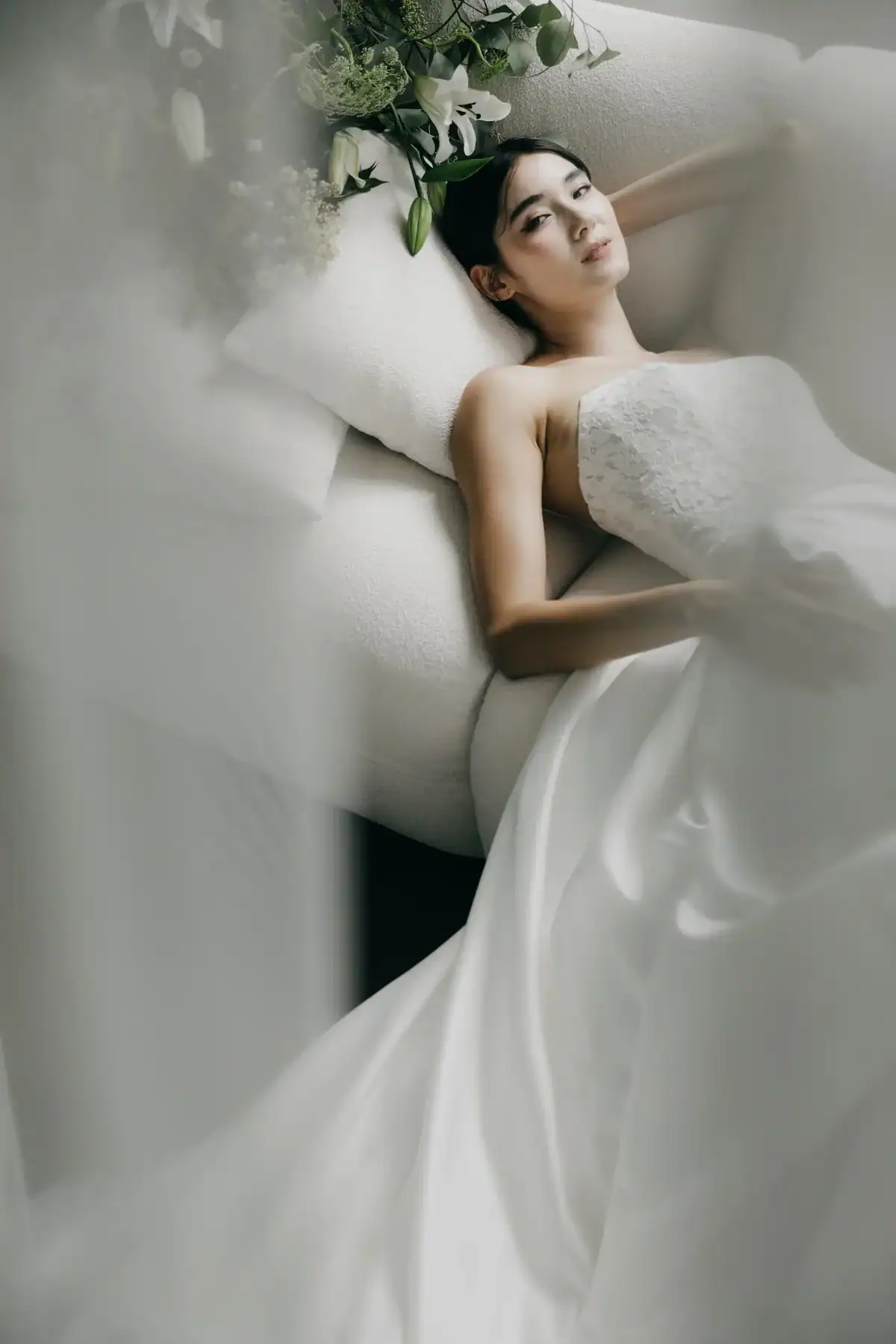 A woman in a strapless white wedding dress lies on a sofa, resting her head on a cushion. Soft, diffused lighting and sheer fabric in the foreground create a dreamy, ethereal atmosphere.