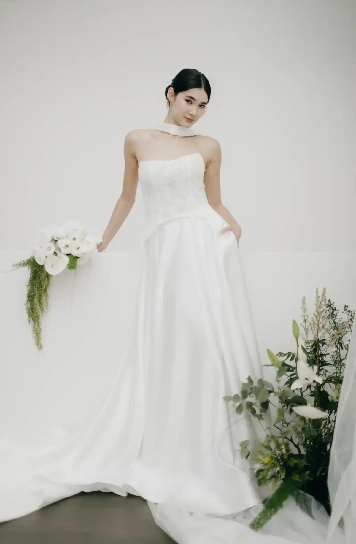 A woman in a strapless white wedding dress holds a bouquet of white flowers and greenery. She stands against a plain white background, with additional flowers and greenery arranged at her feet.
