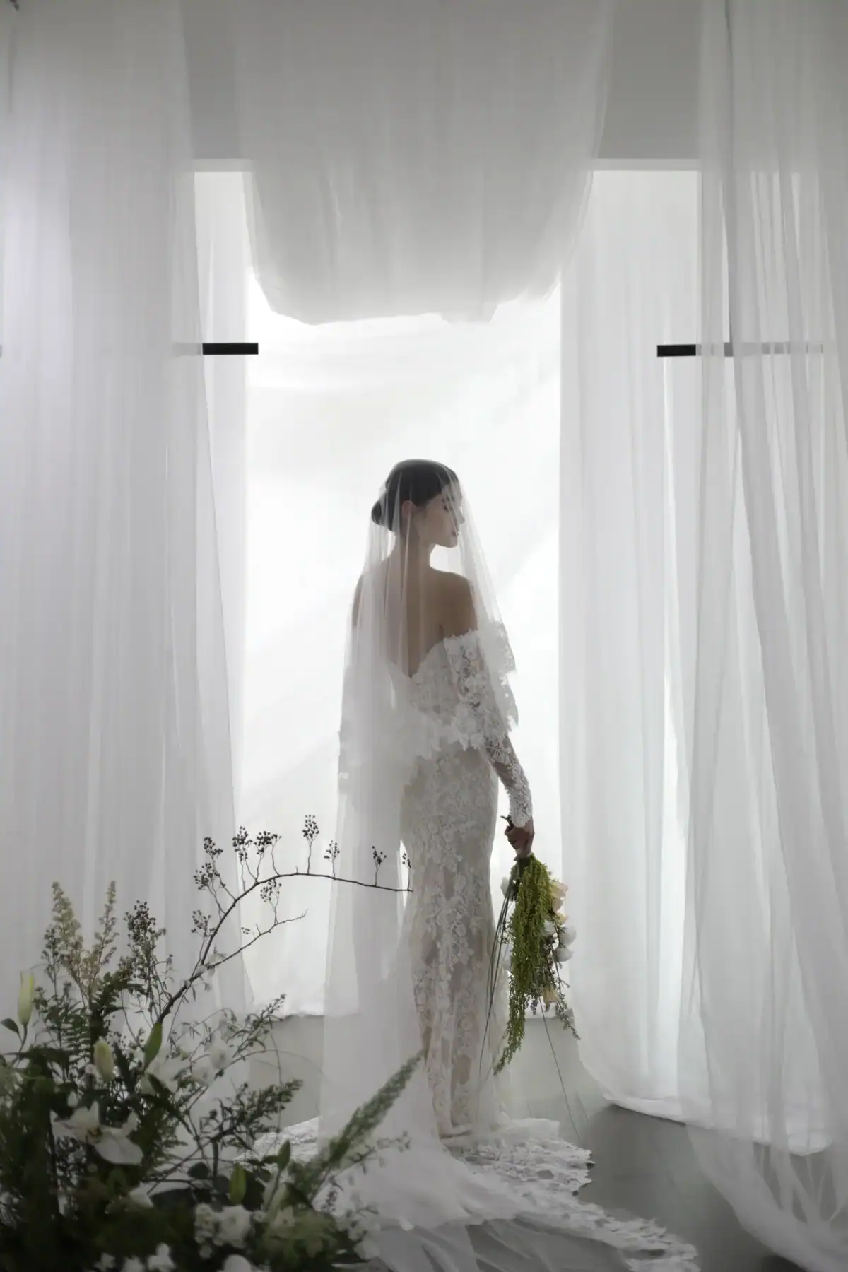 A bride in an off-the-shoulder lace gown and veil stands facing a bright window, holding a bouquet, surrounded by sheer white curtains and delicate floral arrangements.