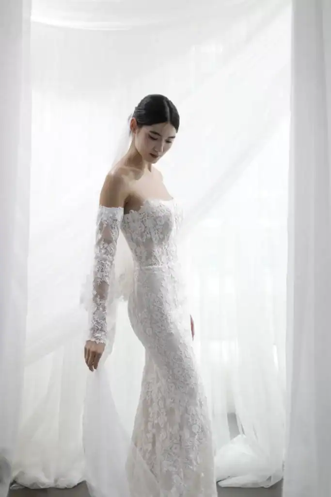 A woman in an elegant, off-the-shoulder lace wedding dress stands gracefully against sheer white curtains, looking down with a gentle expression.