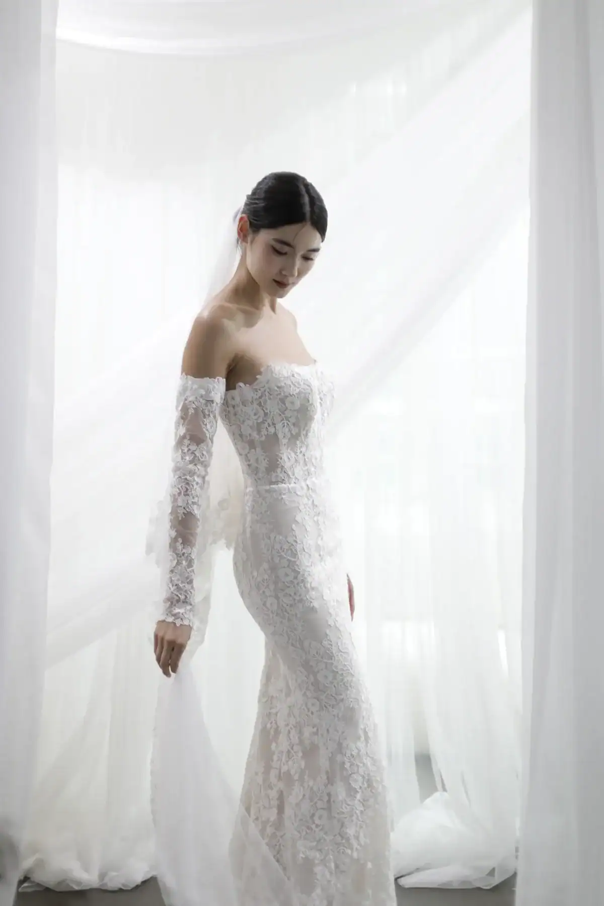 A woman in an elegant, off-the-shoulder lace wedding dress stands gracefully against sheer white curtains, looking down with a gentle expression.