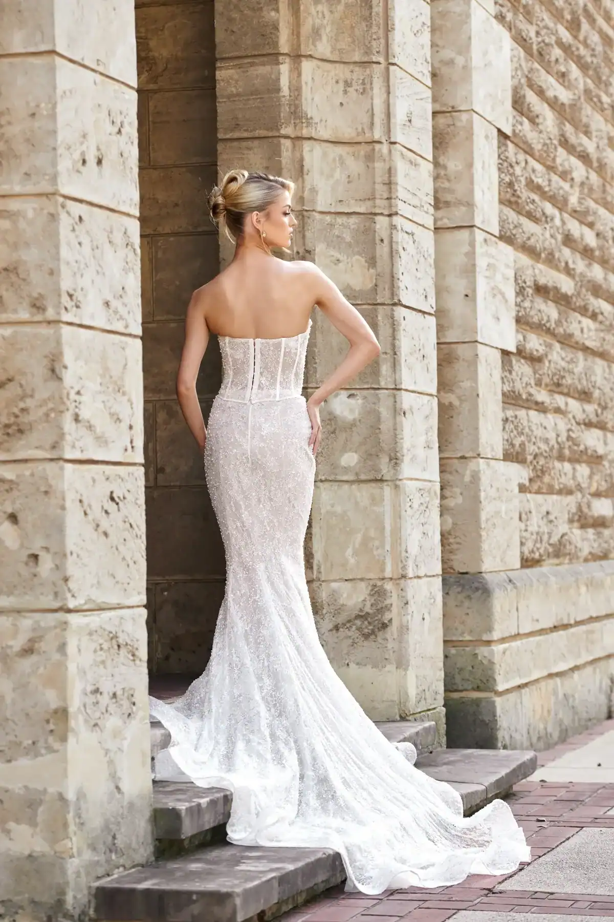 A woman with blonde hair in an updo wears a strapless, figure-hugging white lace gown with a long train, standing on stone steps against a textured stone wall, facing away from the camera.