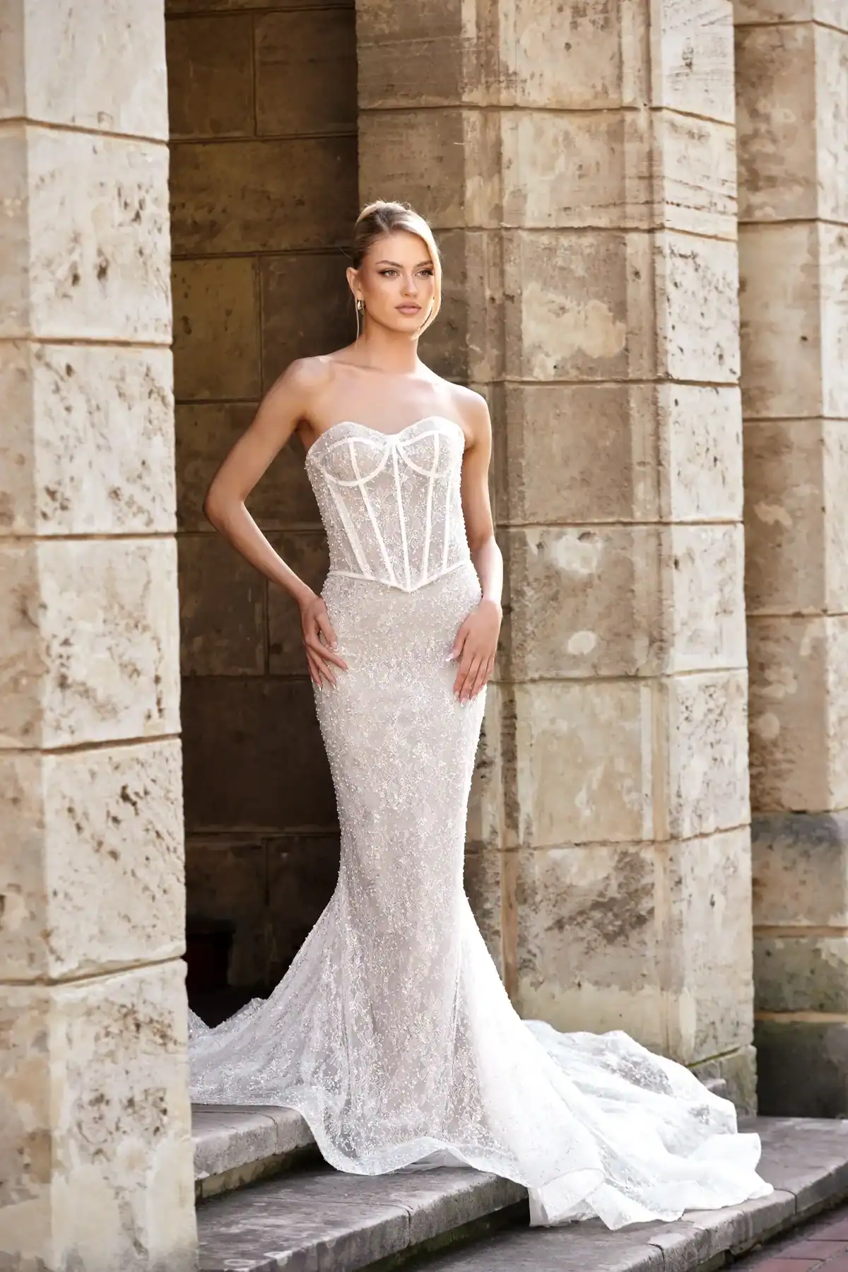 A woman in a strapless, figure-hugging white wedding dress with a structured corset bodice and a long, flowing train stands against a stone column backdrop.
