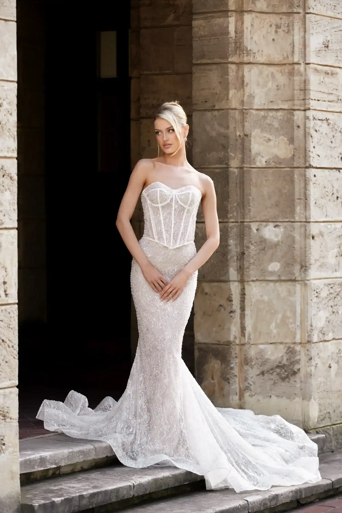 A woman in a strapless, fitted, white lace wedding dress with a structured bodice stands on stone steps in front of a large stone building, hands clasped in front of her.
