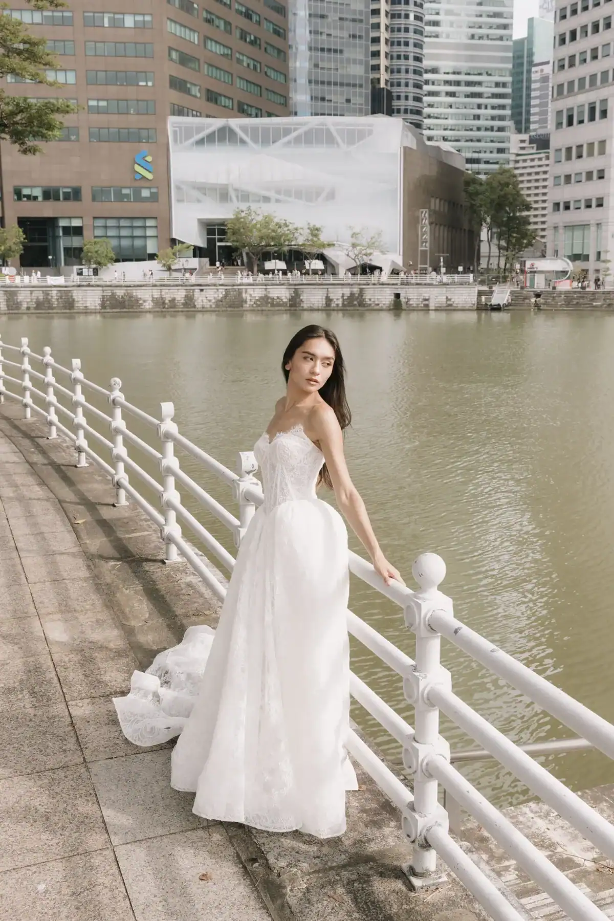 A woman in a white wedding dress stands by a white railing beside a river, with modern city buildings and trees in the background on a bright day.