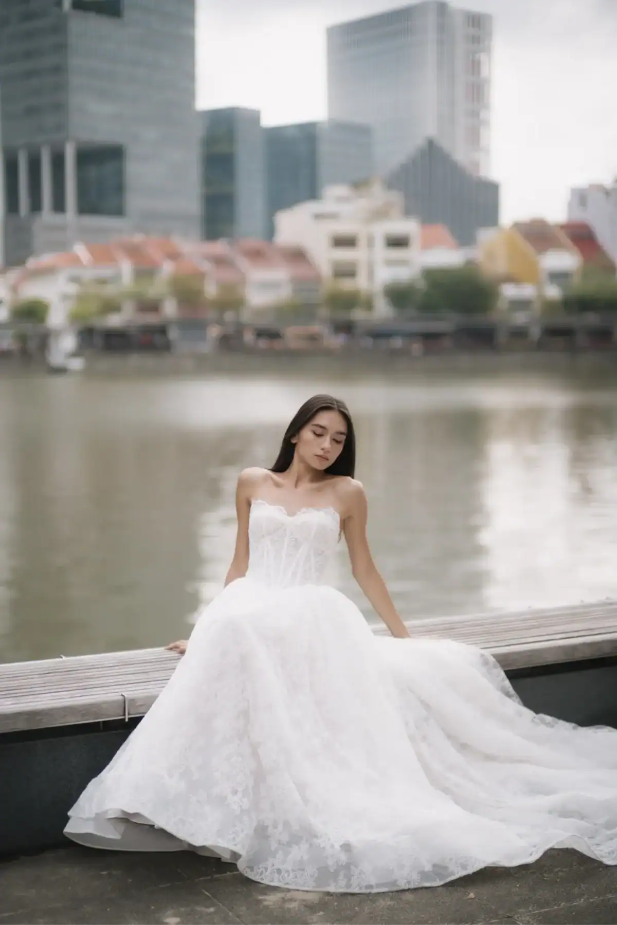 A woman in a white strapless wedding dress sits on a wooden bench by a waterfront, with city buildings, colourful houses, and trees in the background.