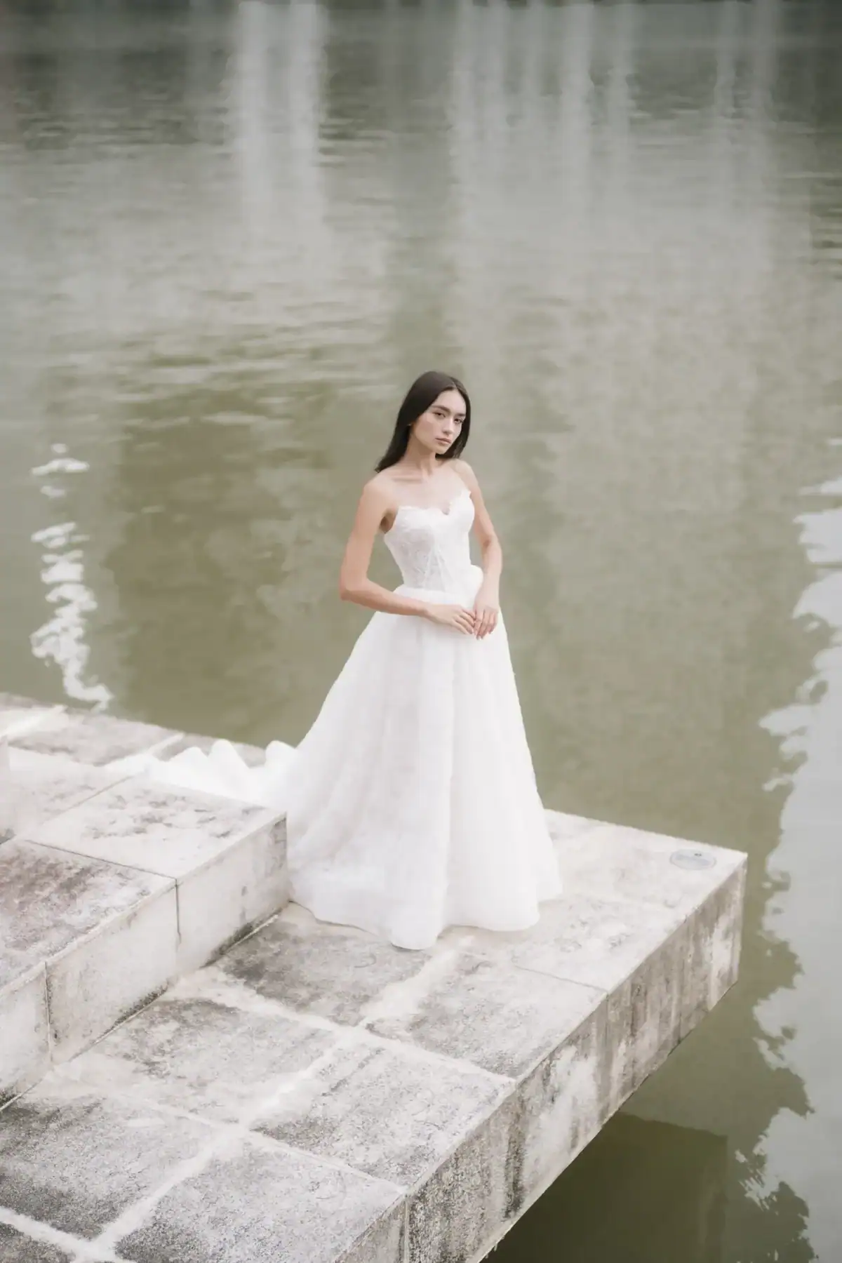 A woman in a white wedding dress stands on stone steps beside a calm body of water, looking slightly to the side with her hands gently clasped in front of her.
