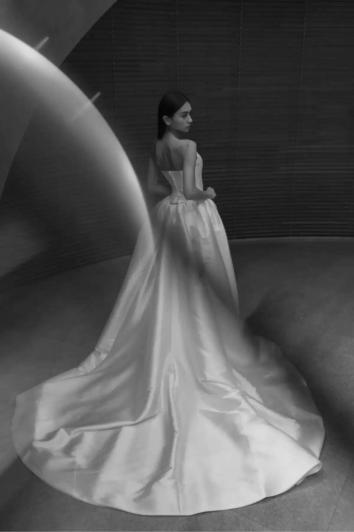 A woman in a strapless, flowing satin gown stands with her back turned, looking over her shoulder. The long train of her dress elegantly spreads out behind her. The image is in black and white with a modern, curved background.