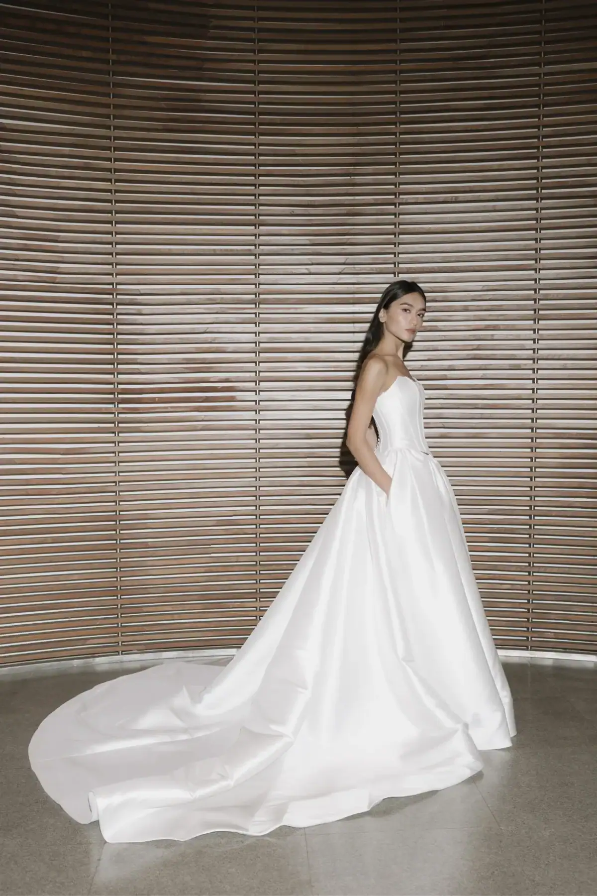 A woman in a strapless, full-skirted white wedding dress stands in front of a modern, wooden slatted wall, looking towards the camera with her hands in the dress’s pockets and the gown’s train trailing behind her.