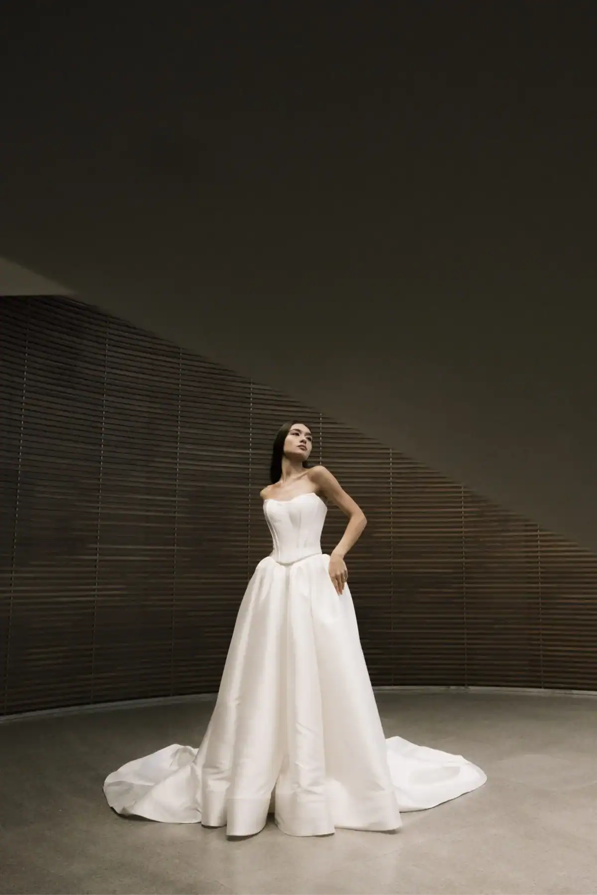A woman in a strapless white ball gown stands indoors on a smooth floor, looking upwards. The background features wood panelling and a geometric ceiling, creating a modern, elegant setting.