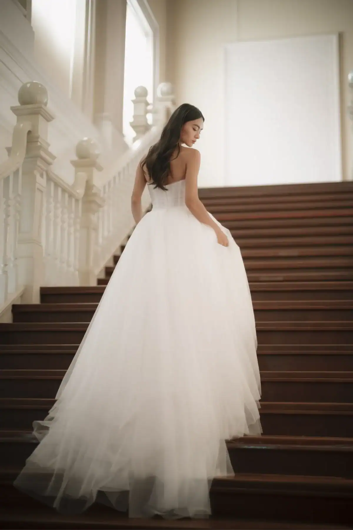 A woman in a strapless white wedding dress with a long tulle train walks up a grand wooden staircase, holding her gown. Soft natural light fills the elegant, spacious setting.