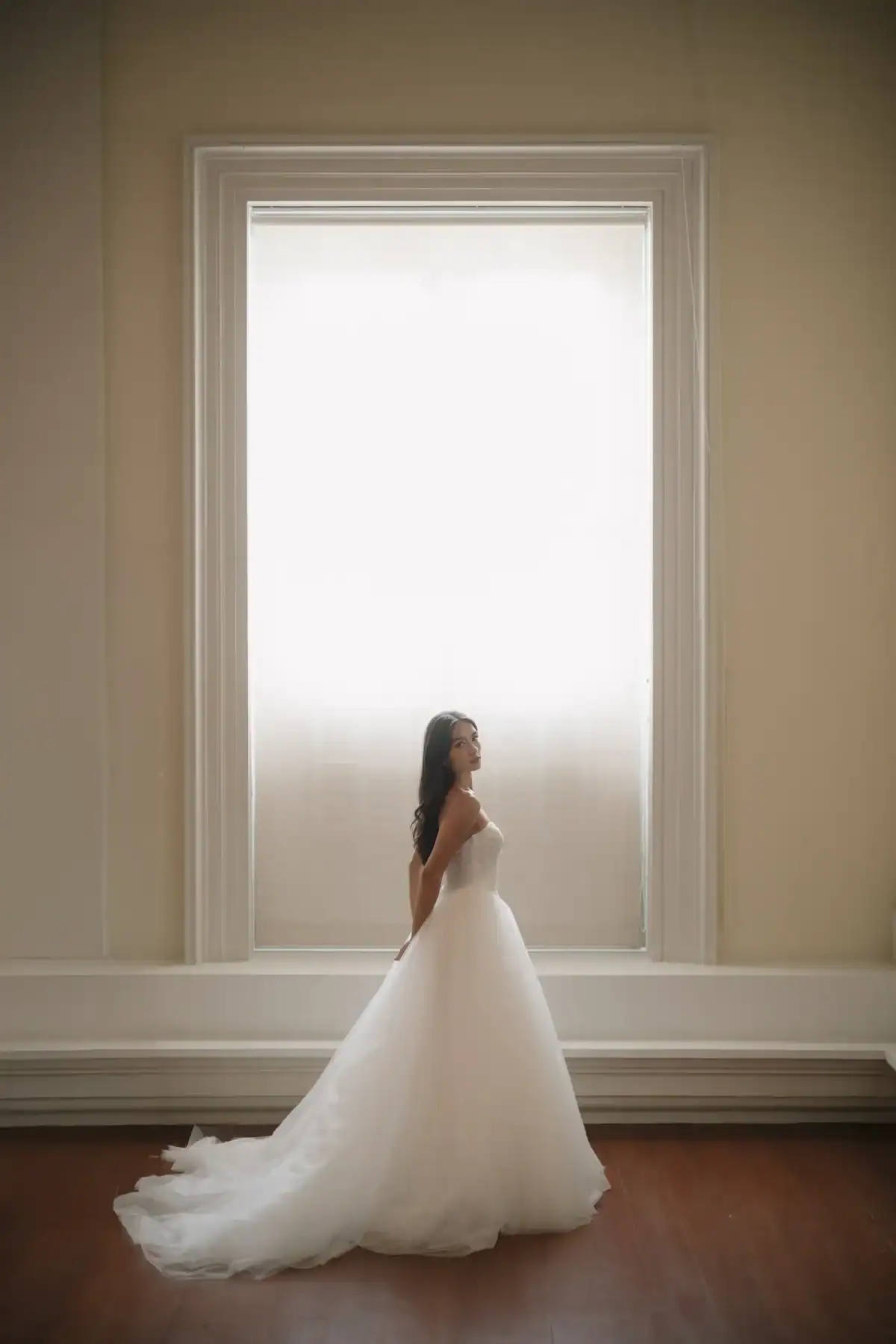 A woman in a white strapless wedding dress stands in front of a large, bright window with soft light streaming in, her hands resting behind her back, and her train flowing onto the wooden floor.