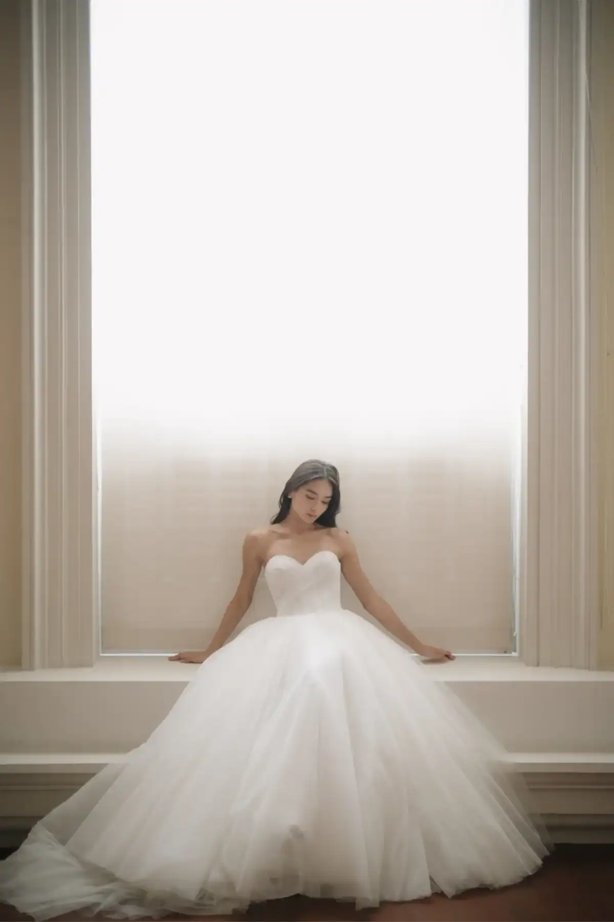 A woman in a strapless white wedding dress sits in front of a large, bright window, looking down with her hands resting on either side of her on the windowsill.