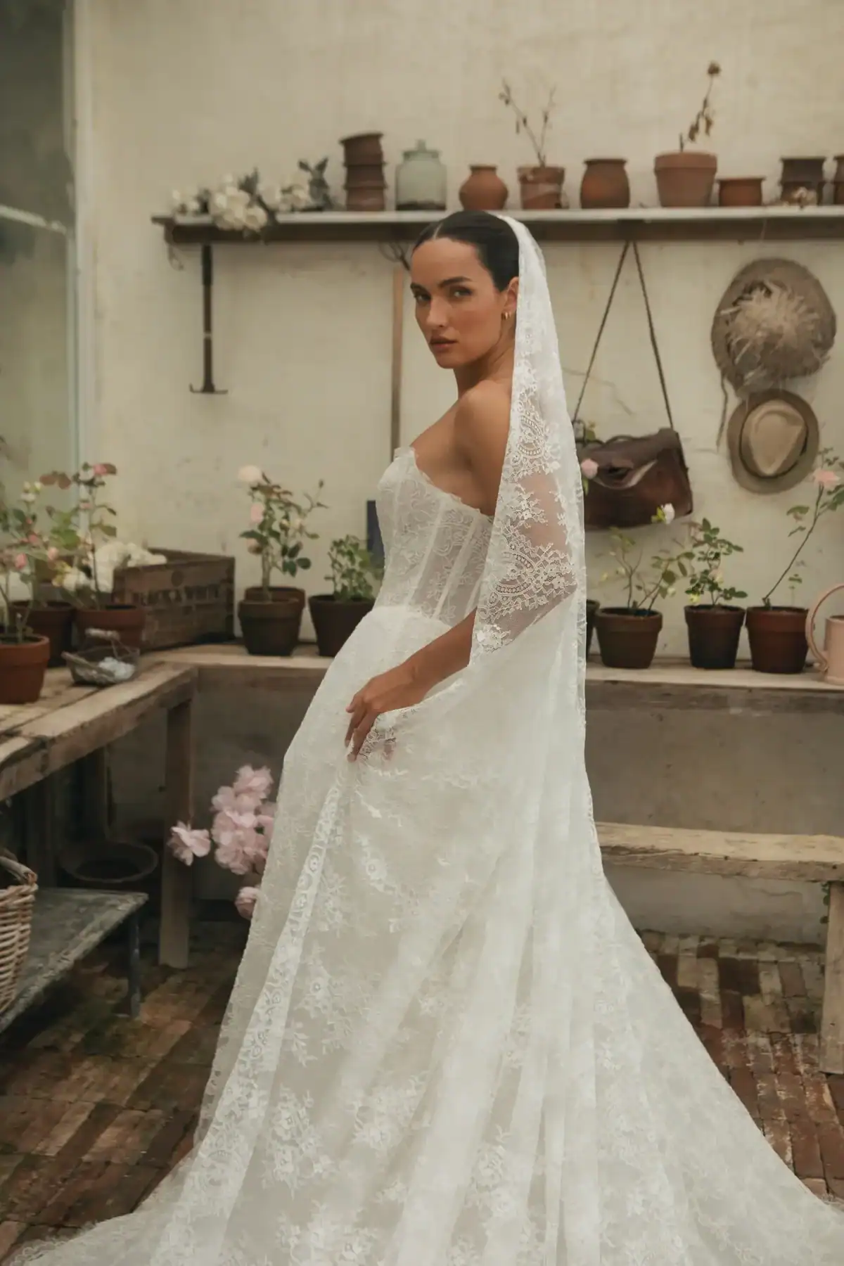 A woman in a strapless lace wedding dress and veil stands in a rustic room with potted plants, wooden shelves, hats, and gardening tools in the background.