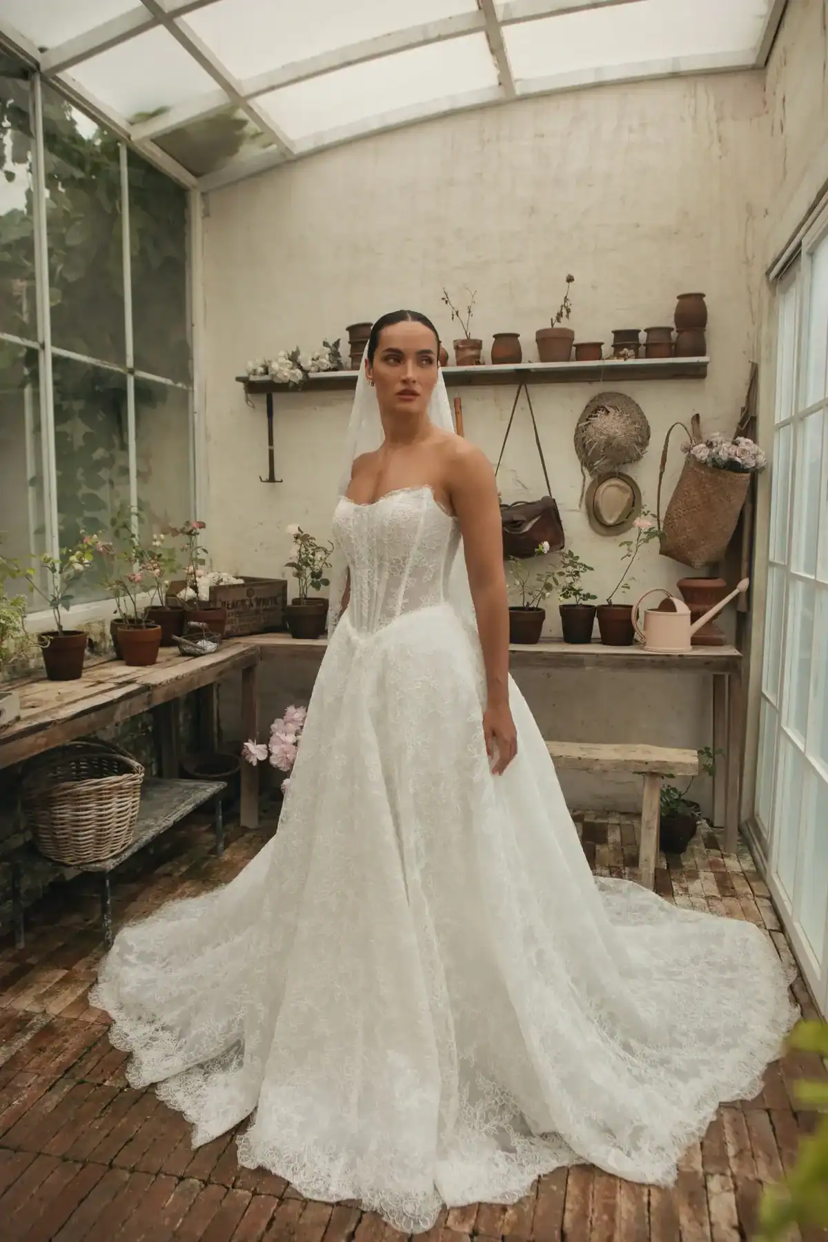 A woman in a strapless white wedding dress and veil stands in a rustic greenhouse with potted plants, garden tools, and wooden shelves in the background.