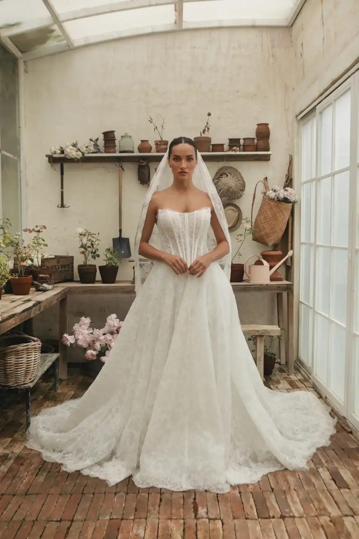A bride in a strapless white lace gown and veil stands in a rustic glasshouse with potted plants, gardening tools, and flowers on shelves and tables around her. Natural light streams in through large glass windows.