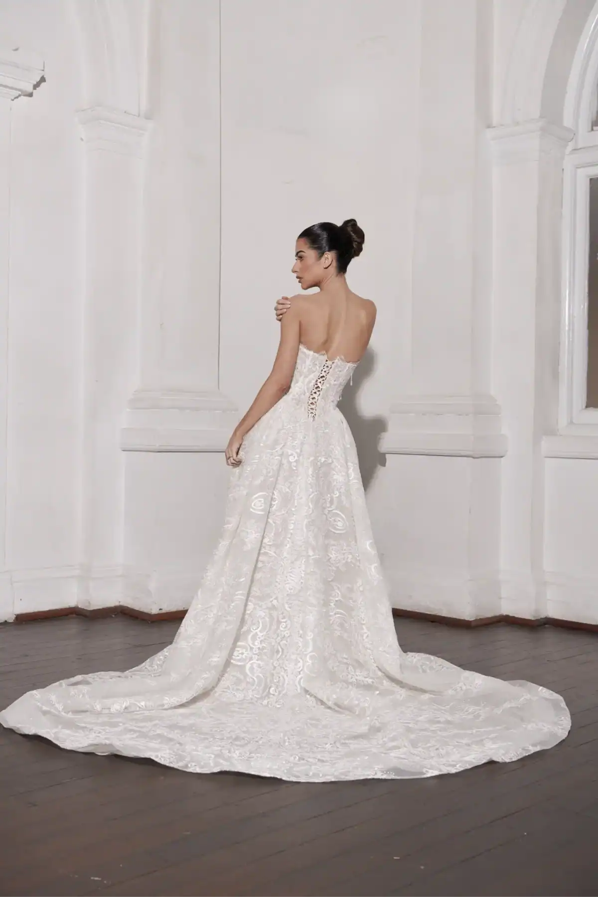 A woman in a strapless, lace, white wedding dress with a long train stands in an elegant white room with high ceilings and arched details, facing away and looking over her shoulder.