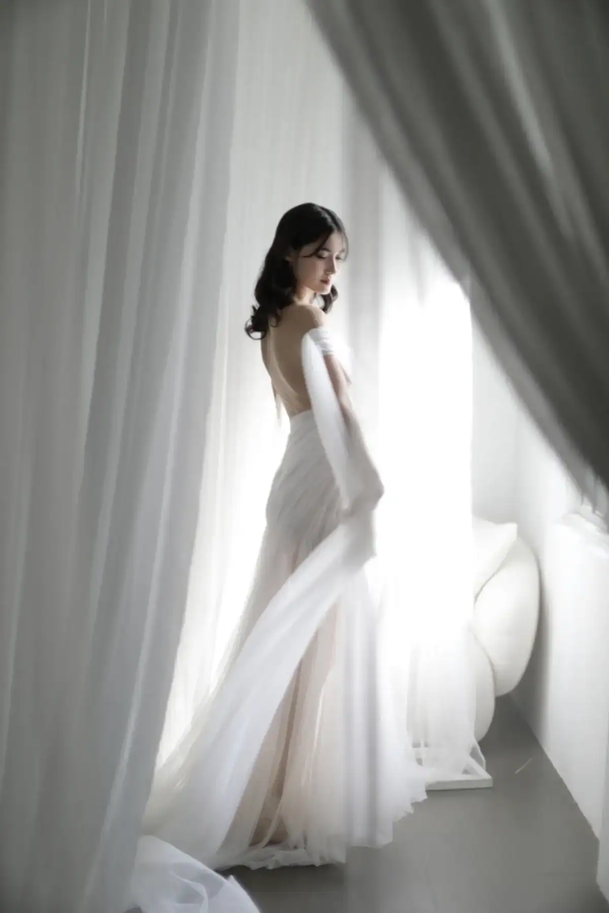 A woman in a flowing, backless white dress stands gracefully between sheer white curtains, bathed in soft natural light from a nearby window.