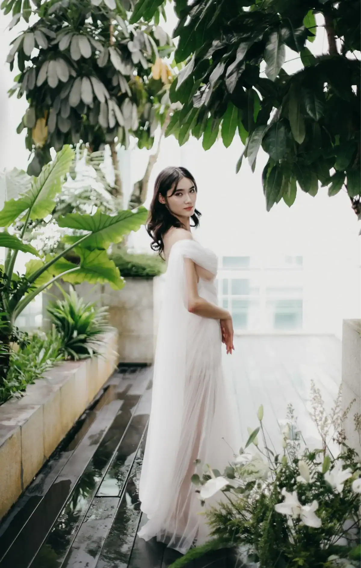 A woman in a flowing white dress stands in a lush indoor garden, surrounded by green plants and trees, looking over her shoulder towards the camera. Natural light fills the bright, serene space.