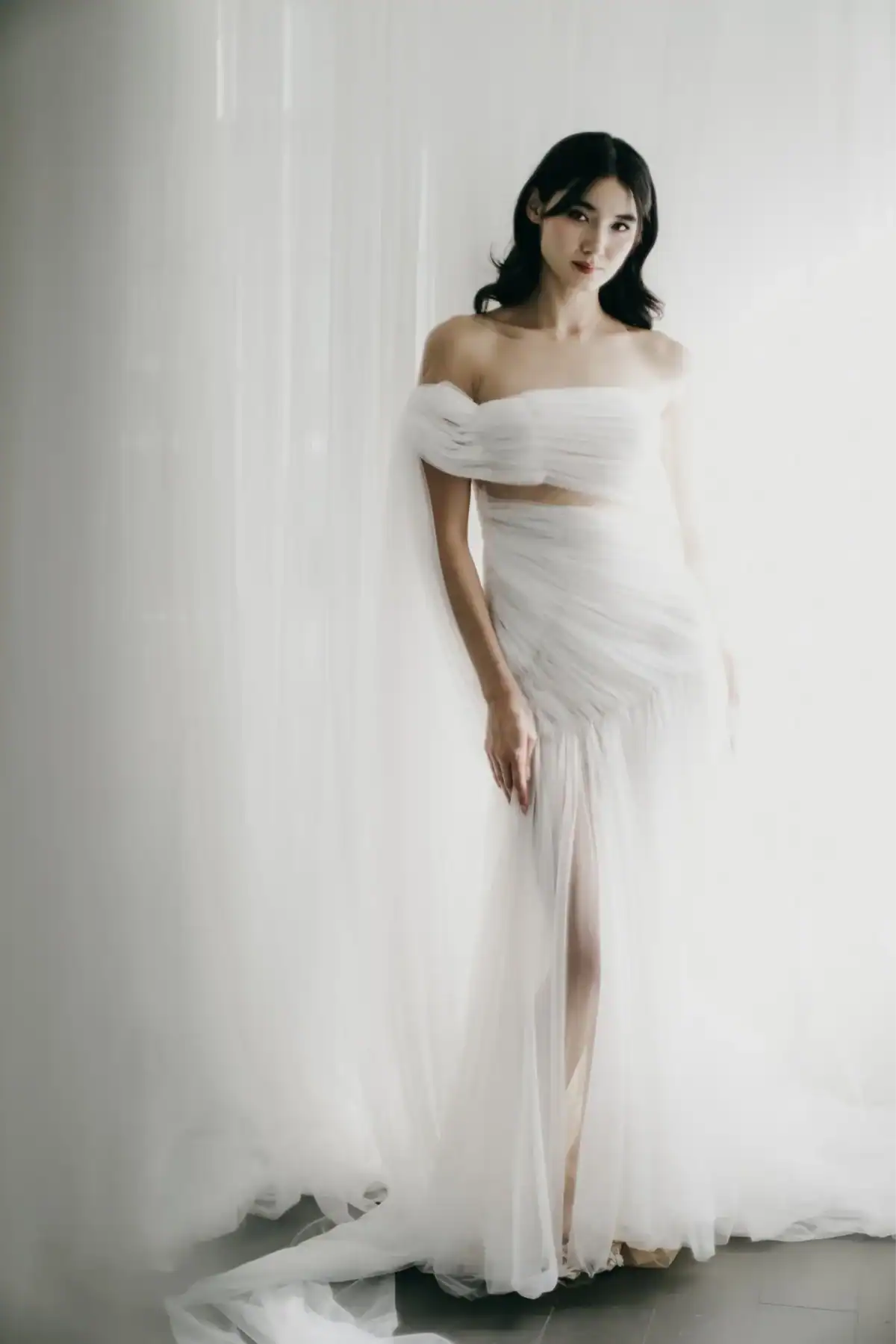 A woman in a sheer, off-the-shoulder white gown stands in soft, natural light against a white curtain backdrop, gazing at the camera with a calm expression.