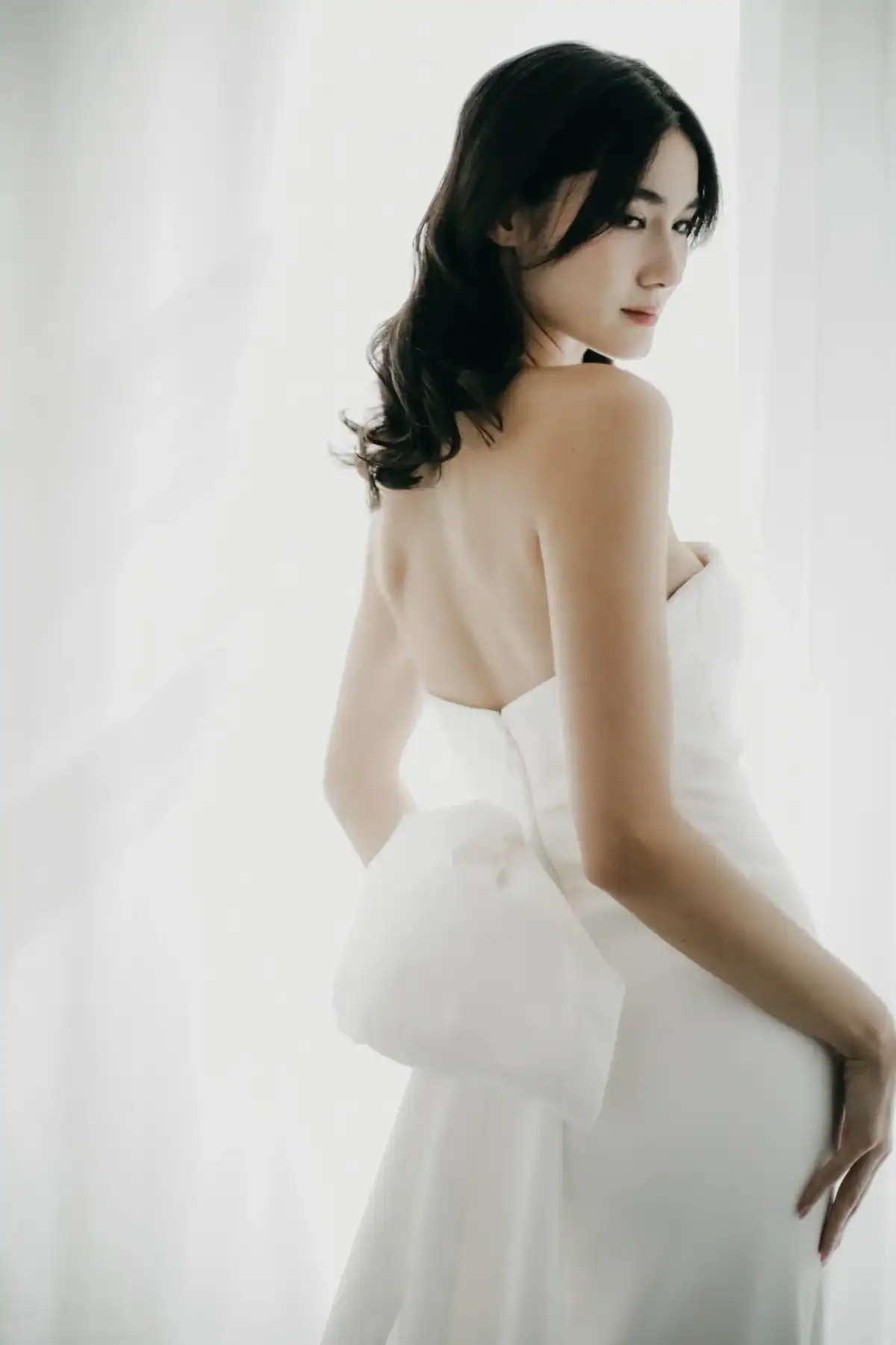 A woman in a strapless white dress with a large bow on the lower back stands against a bright, softly lit background, looking over her shoulder.