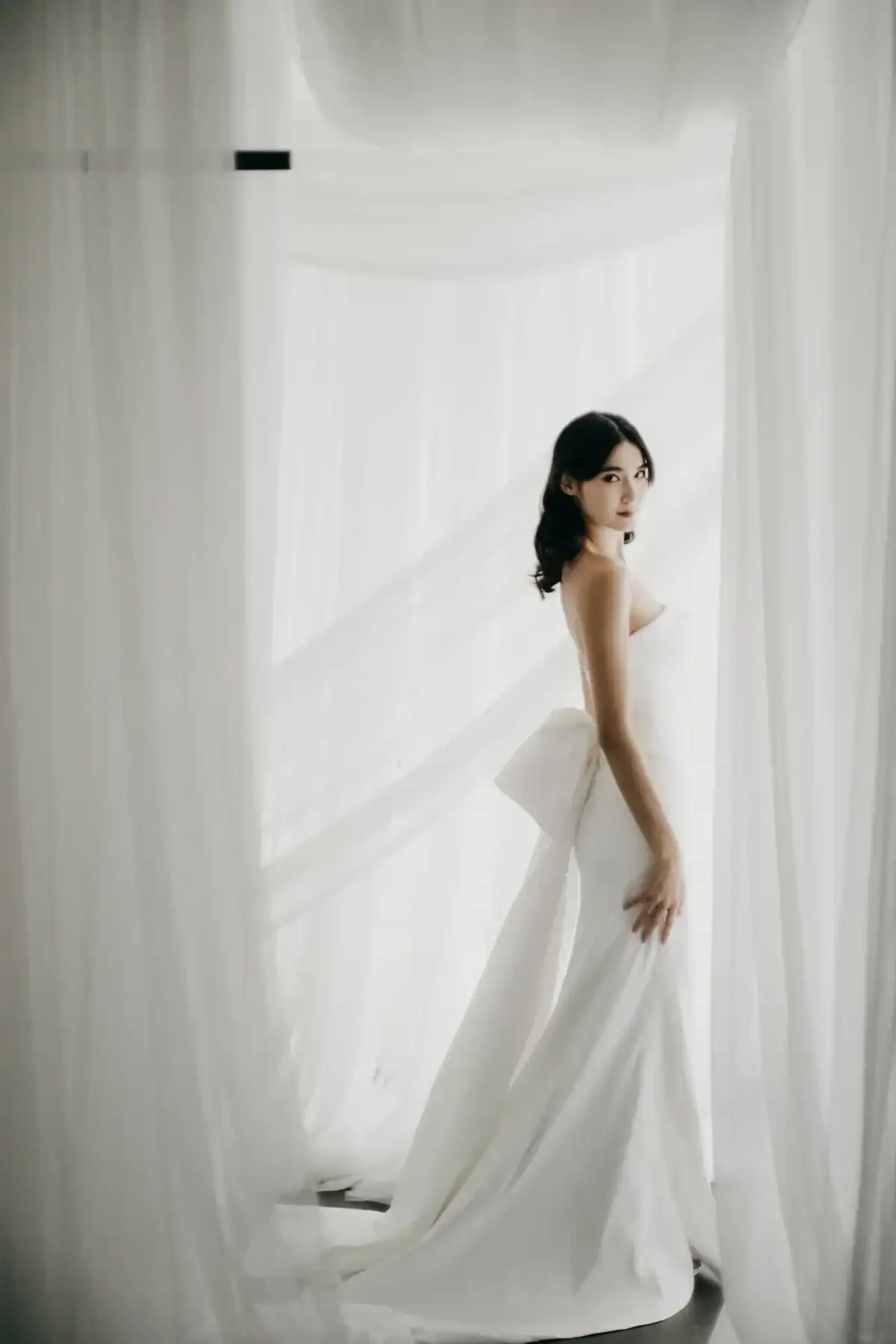 A woman in a strapless white wedding dress with a large bow stands between sheer white curtains, looking over her shoulder in soft, natural light.