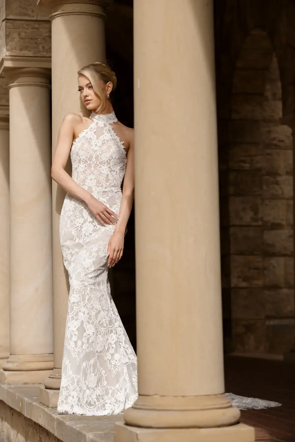 A woman in a fitted, sleeveless white lace gown with a high neckline stands by large stone columns, gazing to the side in an elegant outdoor setting.