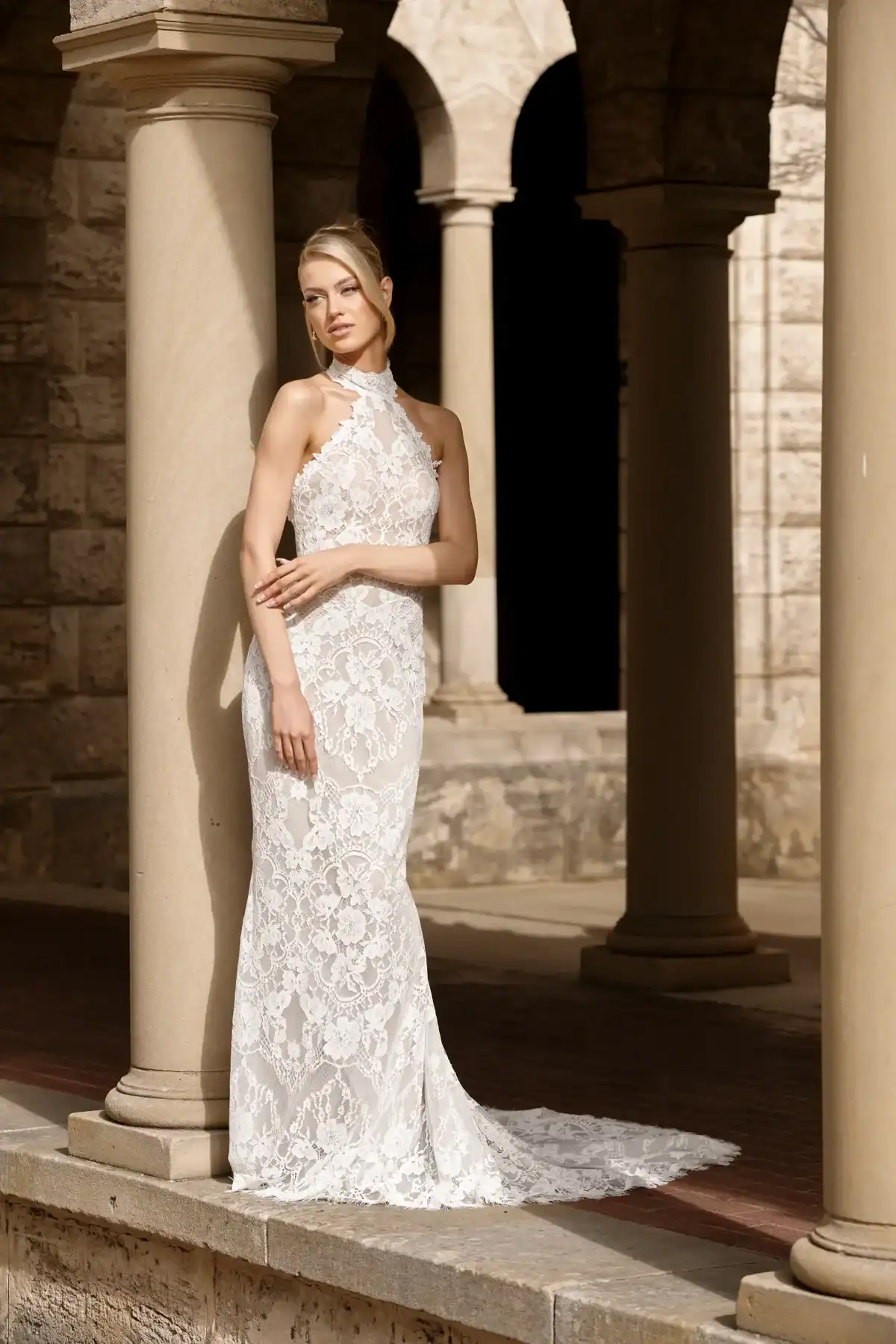 A woman in a sleeveless, high-neck, white lace gown stands against a stone column in an outdoor archway, with stone walls and pillars in the background.