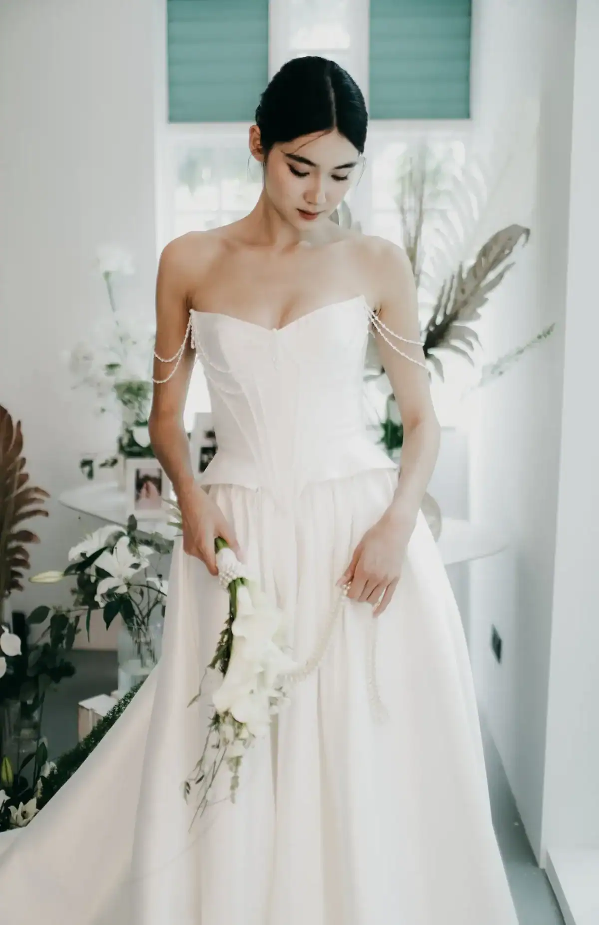 A bride in a strapless white gown with pearl details holds a bouquet of white flowers, standing indoors surrounded by green plants and soft natural light.