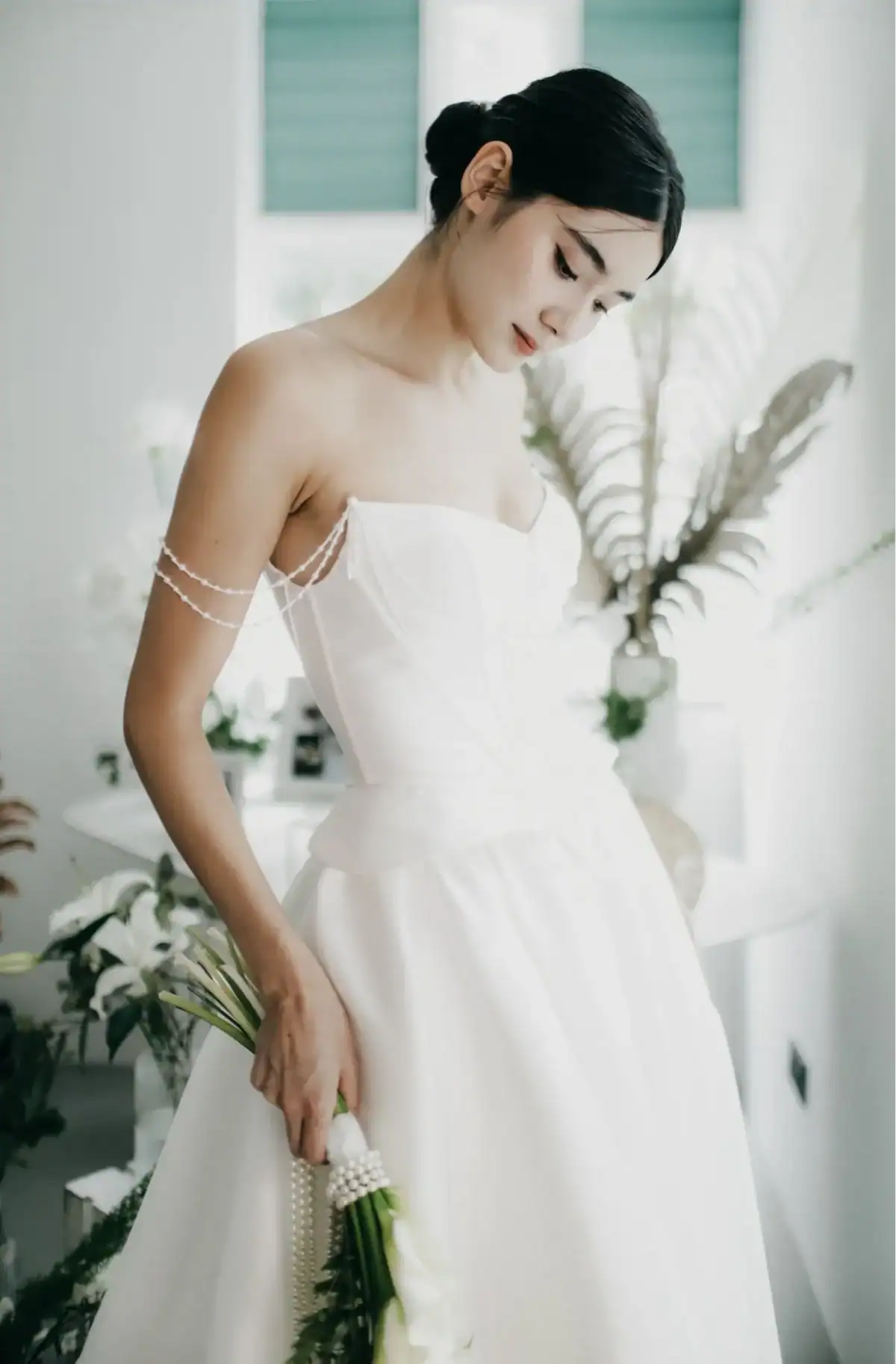 A woman in a white off-shoulder wedding dress holding a bouquet of flowers stands indoors, surrounded by greenery and soft natural light, looking downwards with a gentle expression.