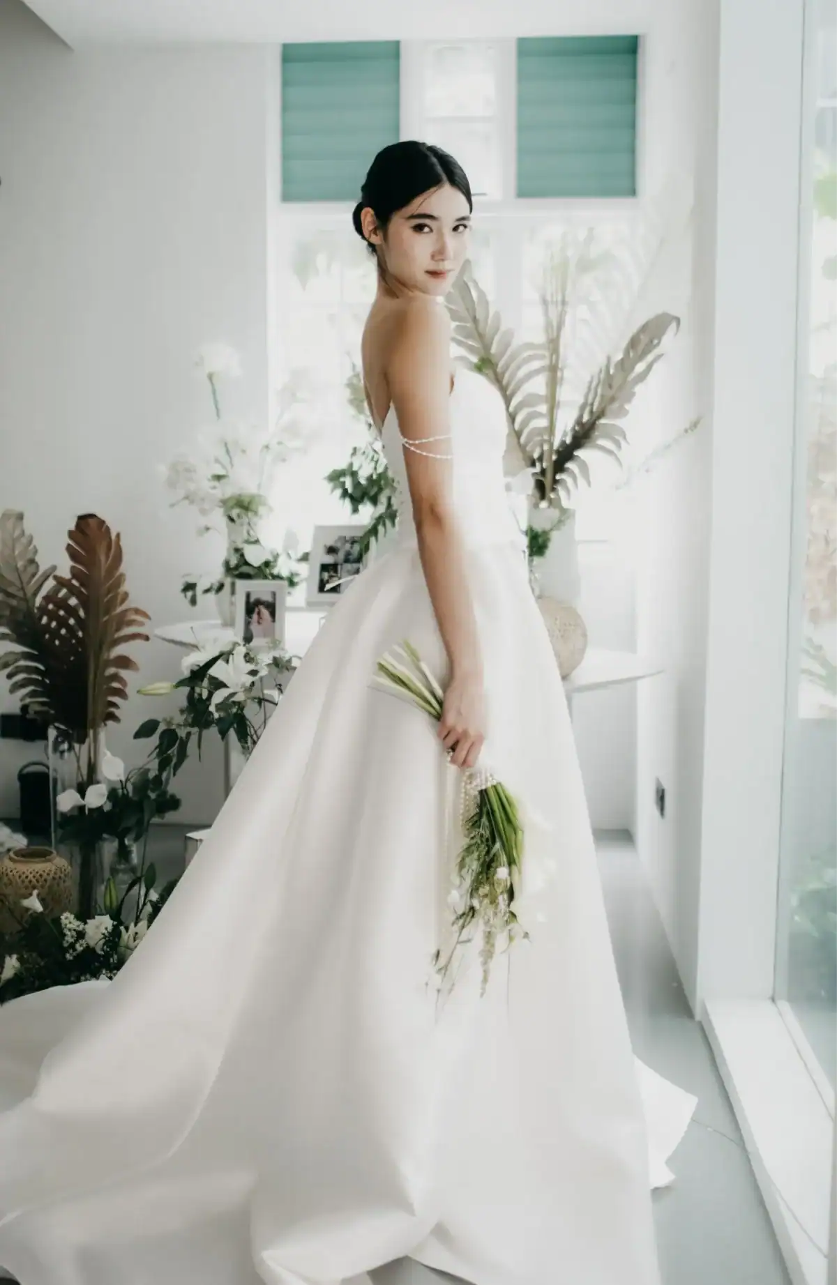 A woman in an elegant white wedding dress stands indoors, holding a bouquet of white flowers. She looks over her shoulder, surrounded by greenery and natural light from large windows.