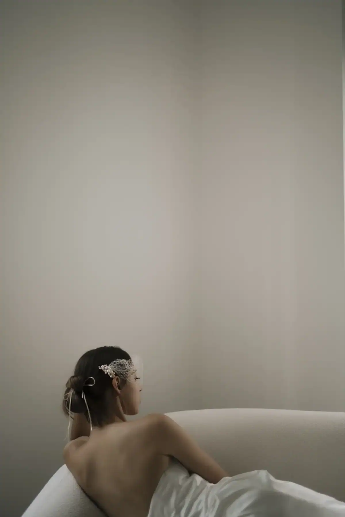 A woman with her back facing the camera sits in a bath, wearing a decorative hairpiece and a white mask. The neutral-toned background and minimal decor create a calm, serene atmosphere.