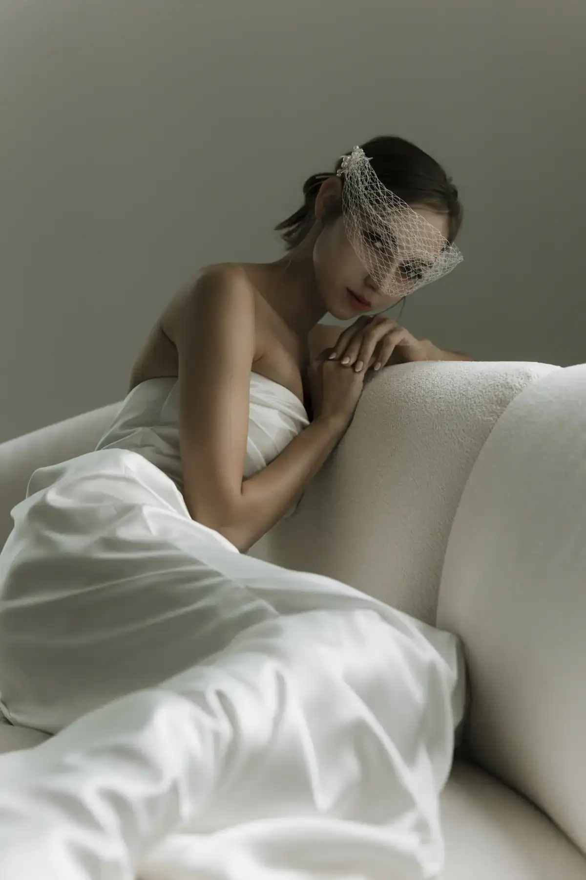 A woman in a strapless white dress reclines on a light-coloured sofa, wearing a delicate birdcage veil over her face. She gazes softly at the camera in a minimalist setting.