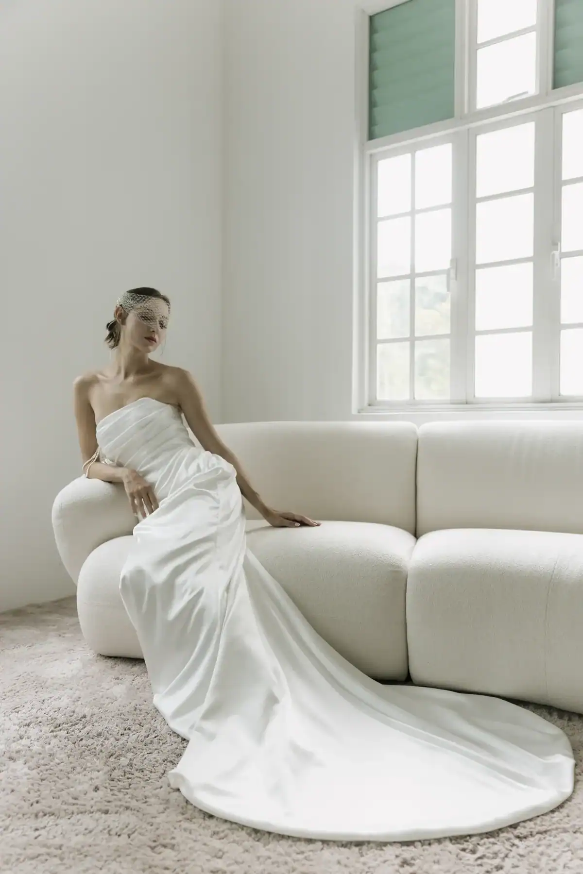 A woman in a strapless white wedding dress with a long train and a birdcage veil sits gracefully on a cream-coloured sofa in a bright, minimalist room with large windows.