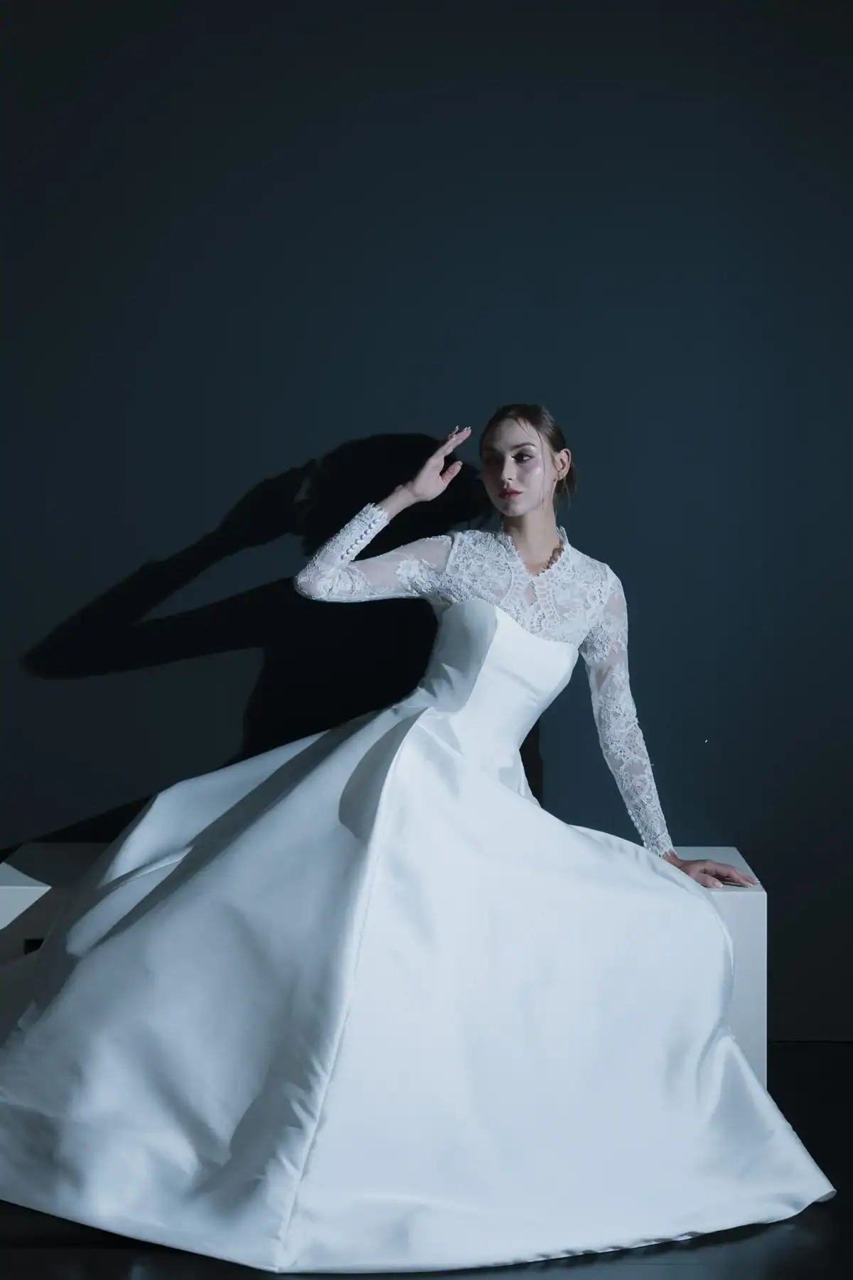 A woman in a long white wedding dress with lace sleeves sits gracefully on a white block against a dark background, casting a shadow on the wall behind her.