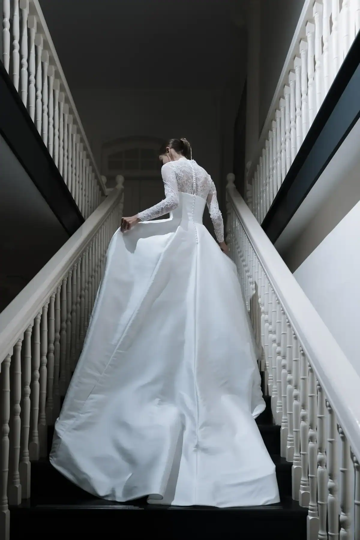 A bride in a long white gown with lace sleeves ascends a staircase, holding up her dress. The view is from behind, highlighting the flowing train and elegant details of her wedding dress.