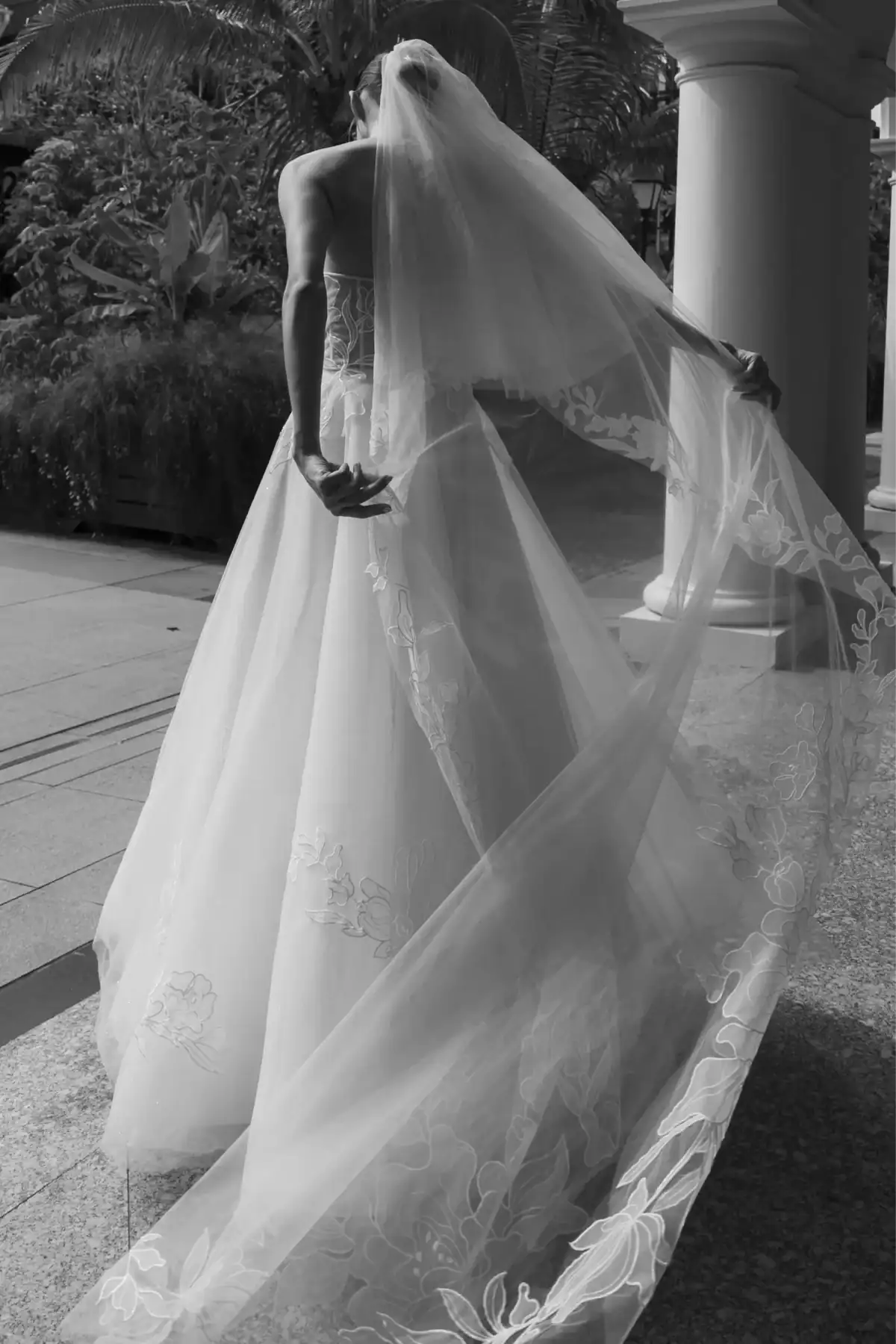 A bride in a flowing, embroidered wedding dress and veil stands outdoors, holding her veil with one hand as it cascades behind her. The scene is elegant and sunlit, surrounded by greenery and architectural columns.